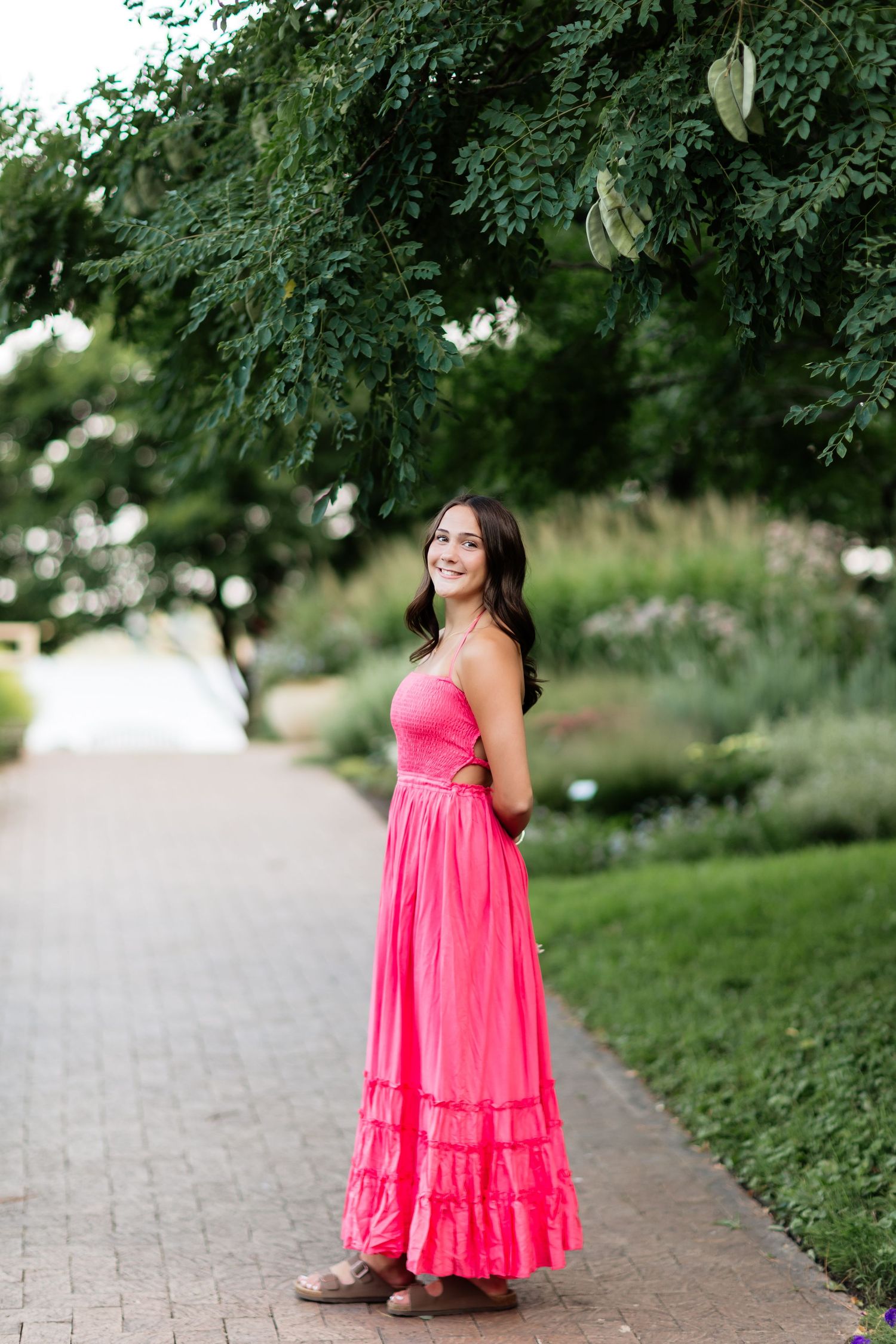 Young woman in bright pink strapless tiered maxi dress standing on a brick path in a lush garden.