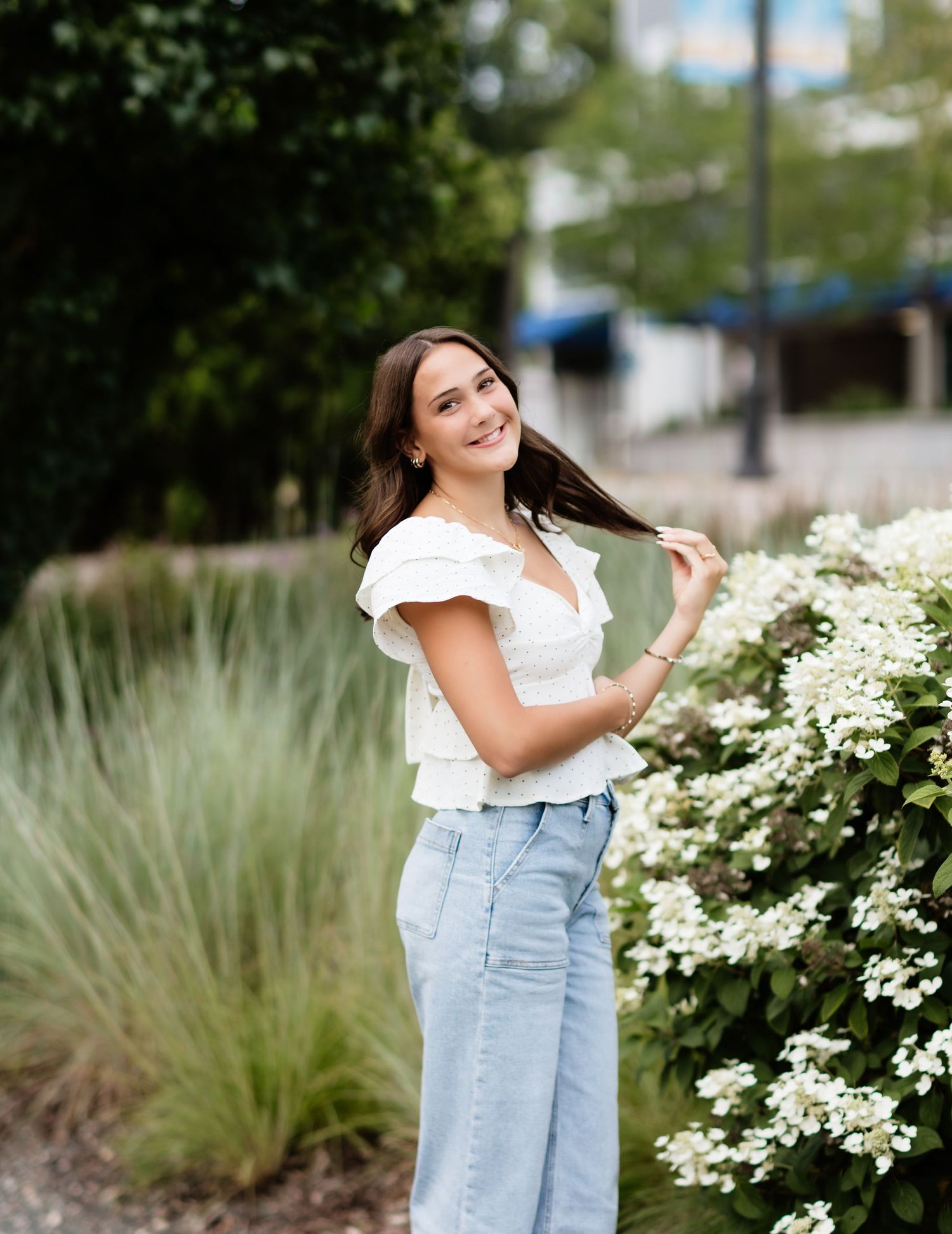 Young woman in white top and jeans smiling among white hydrangea blooms in a lush garden setting.
