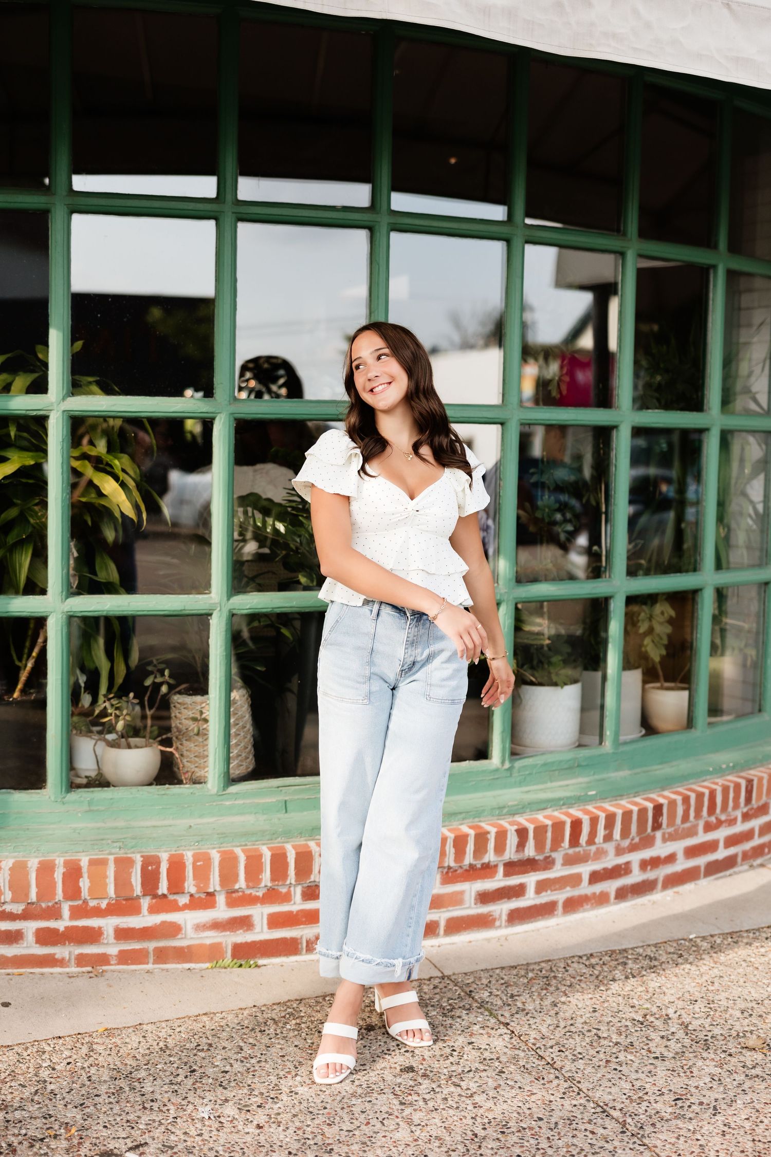 Young woman in white ruffle top and light jeans smiling outside green brick-trimmed storefront.