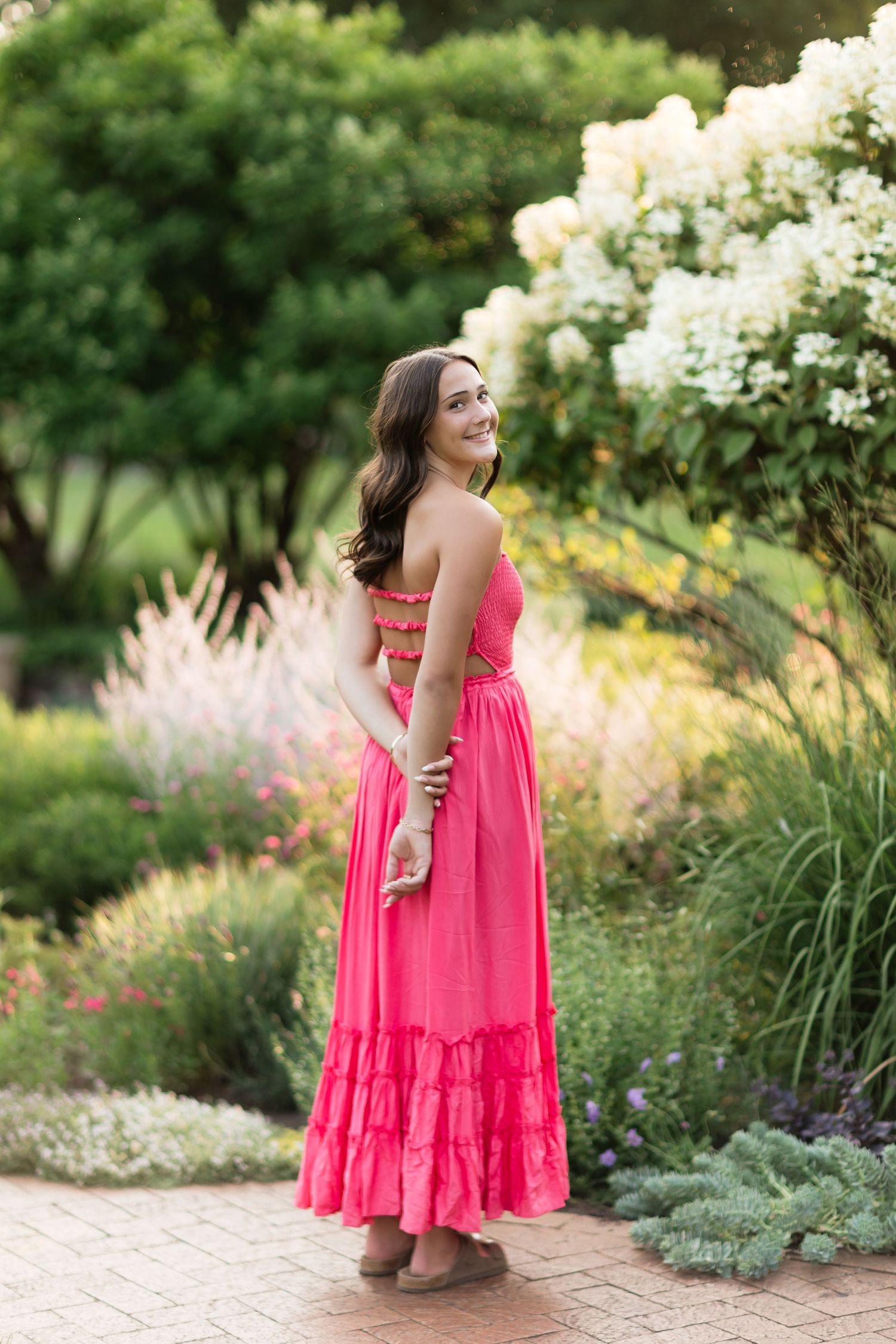 Woman in pink maxi dress smiling in a lush garden with white flowers.