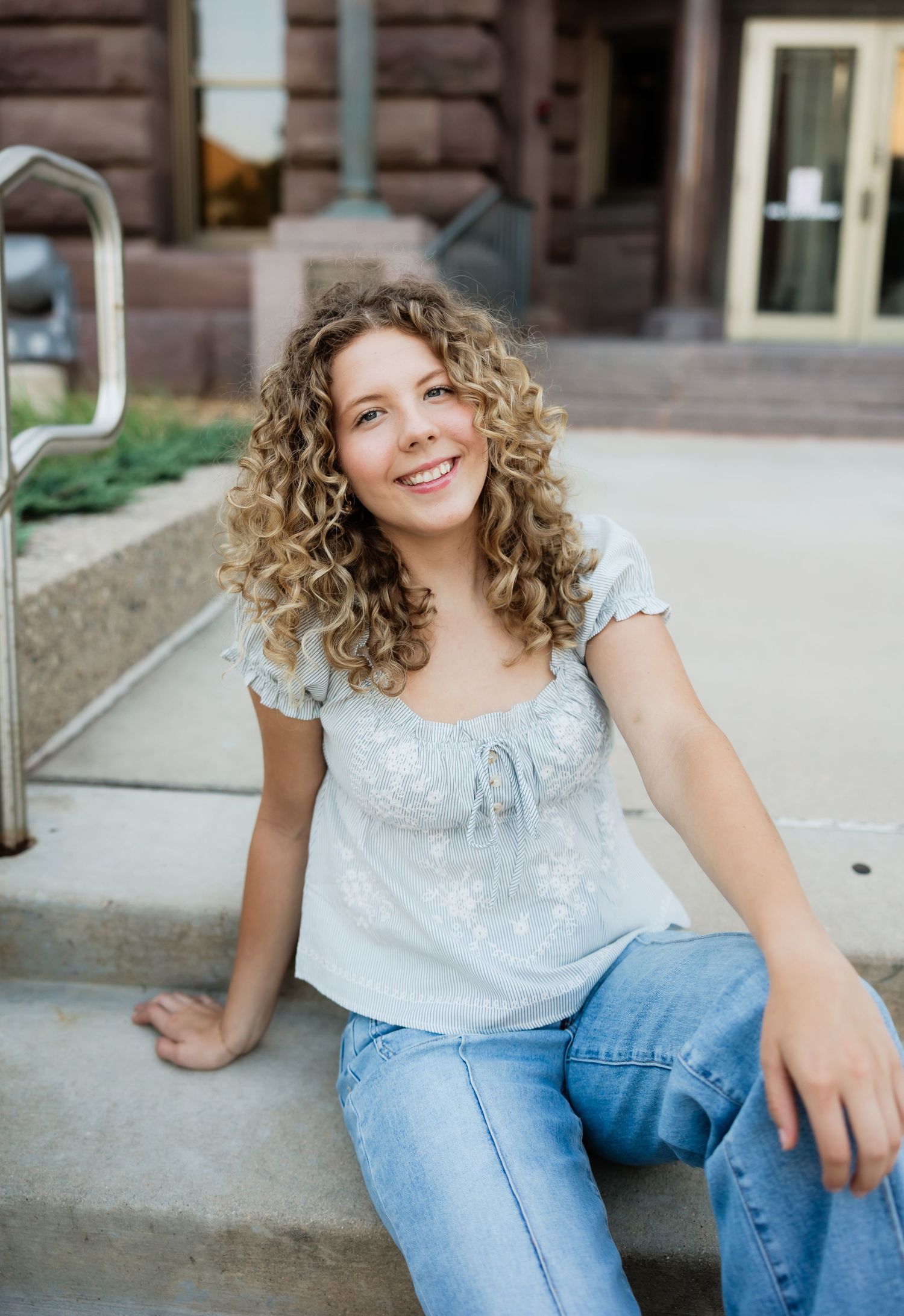 Smiling curly-haired teen in light blue top and jeans sitting on outdoor steps near a building.
