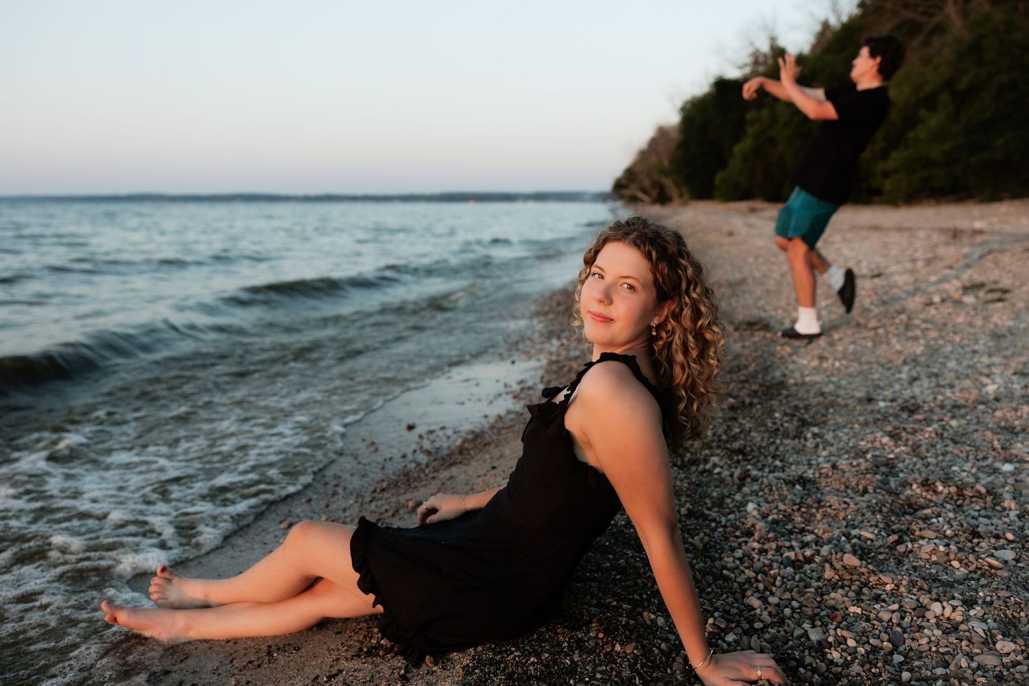 Young woman in black dress sitting on a pebble beach at dusk, with a person running in the background.