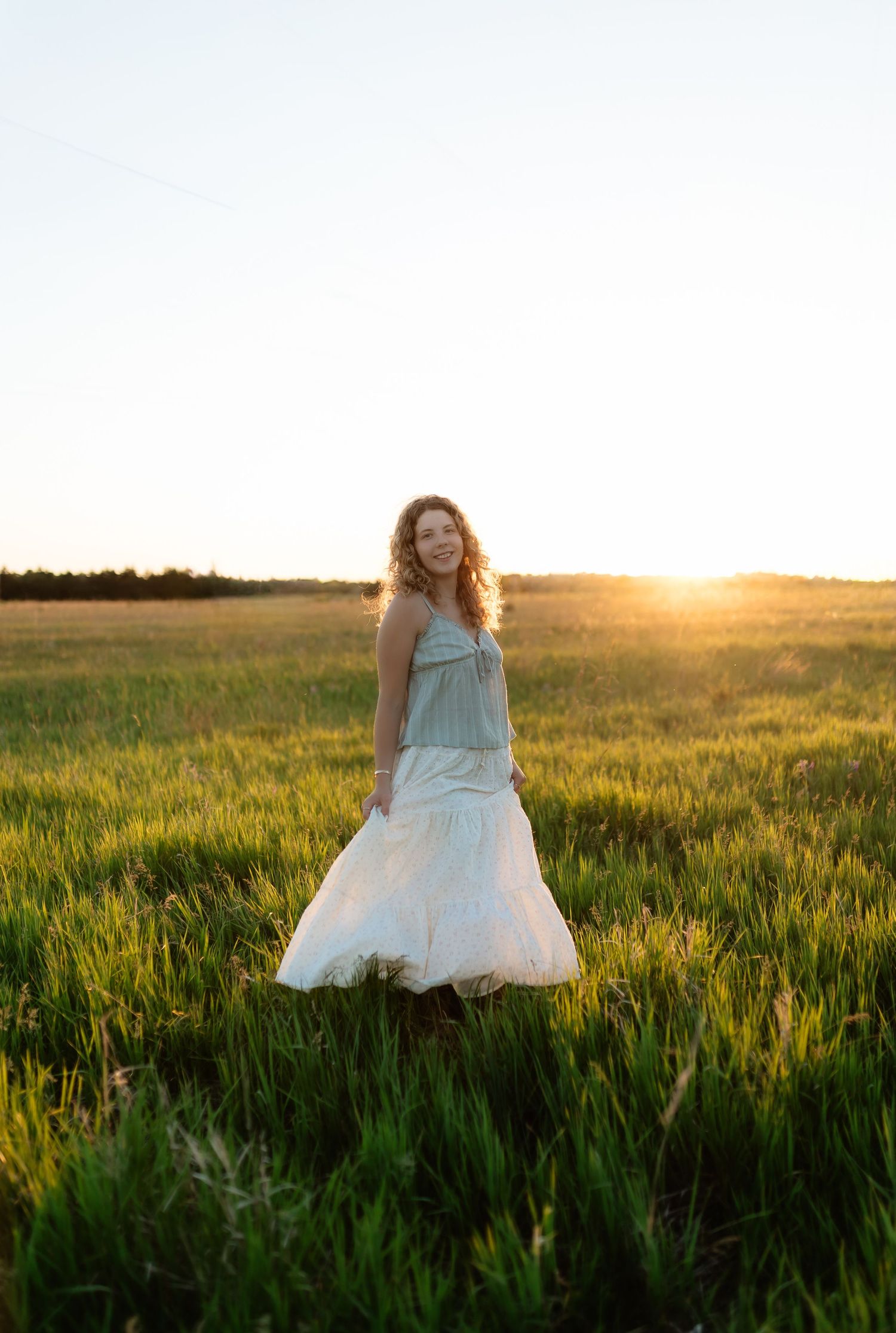 Young woman in mint and white dress twirling in open green field during stunning golden sunset.