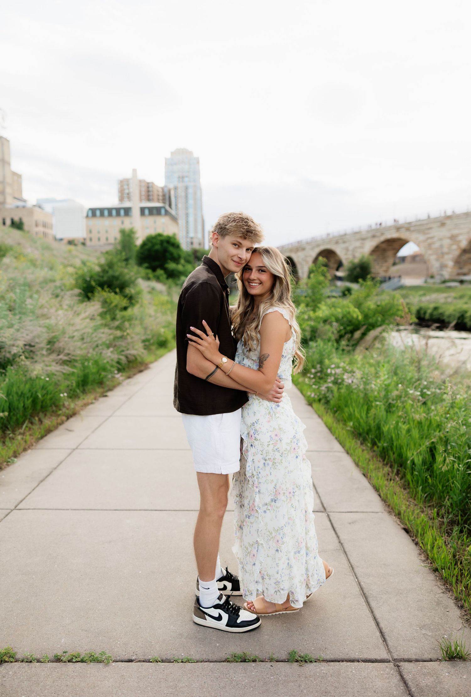 Smiling couple posing together on a scenic Minneapolis riverfront path with the Stone Arch Bridge behind them.