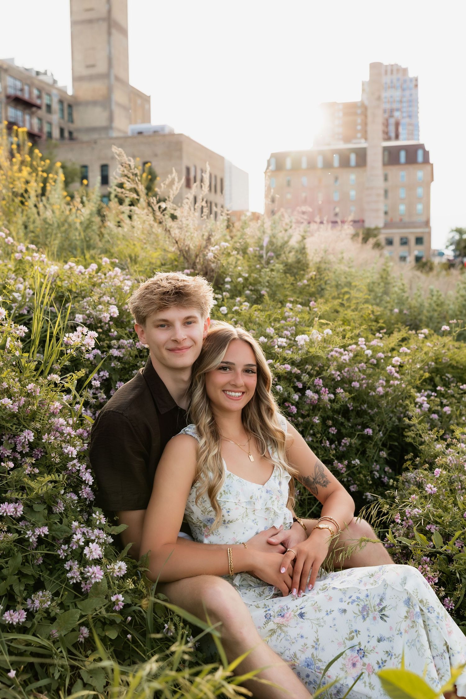 Smiling couple sitting together in a lush wildflower field with golden sunlight and city buildings behind them.