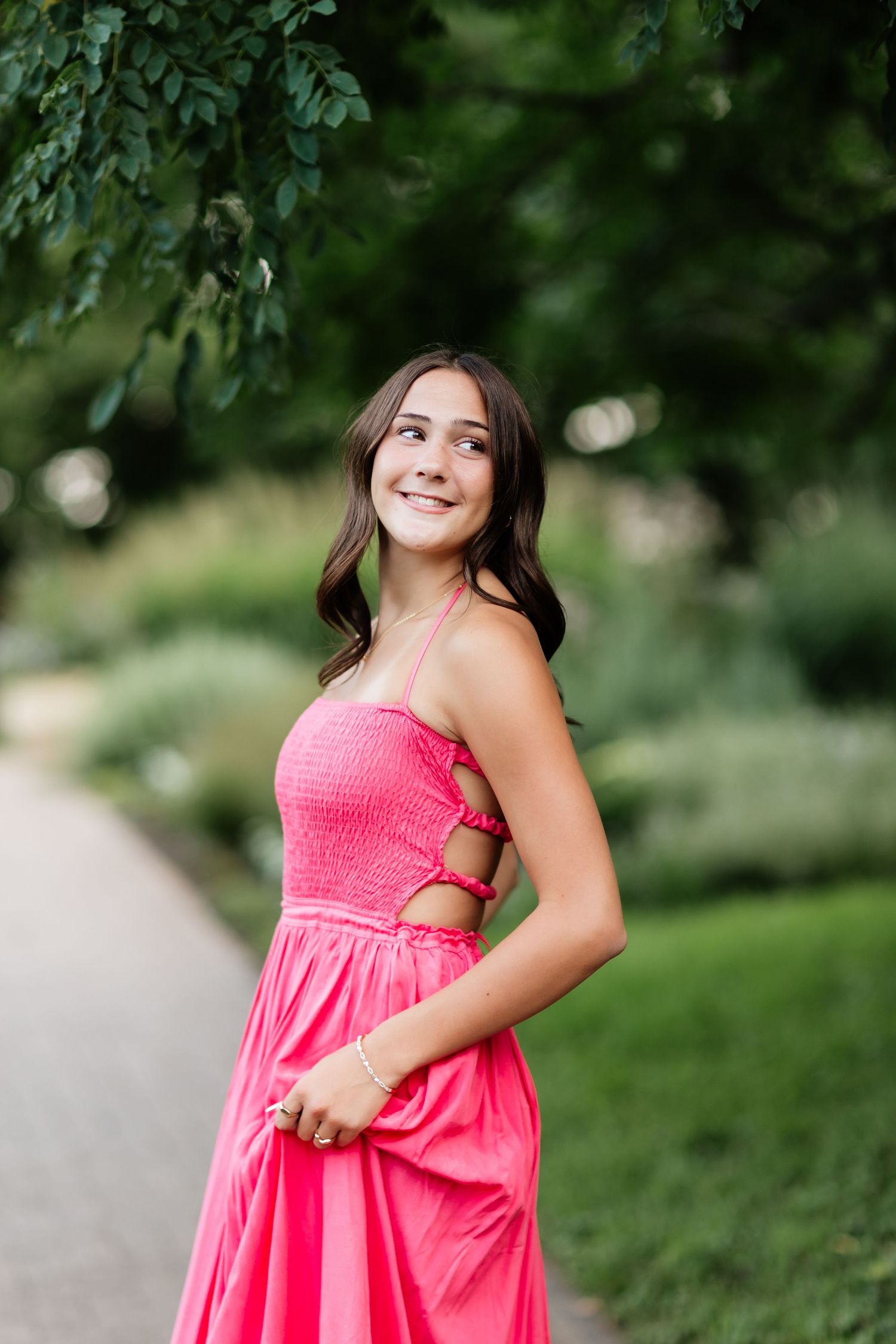 Smiling young woman in bright pink halter dress posing outdoors along a lush green garden path.