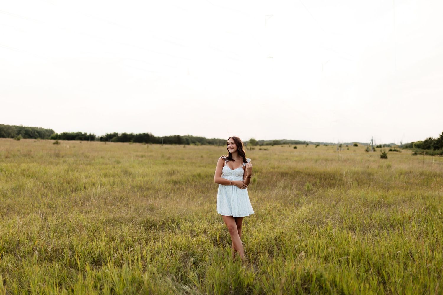 Young woman in white dress standing alone in wide open grassy field under overcast sky.
