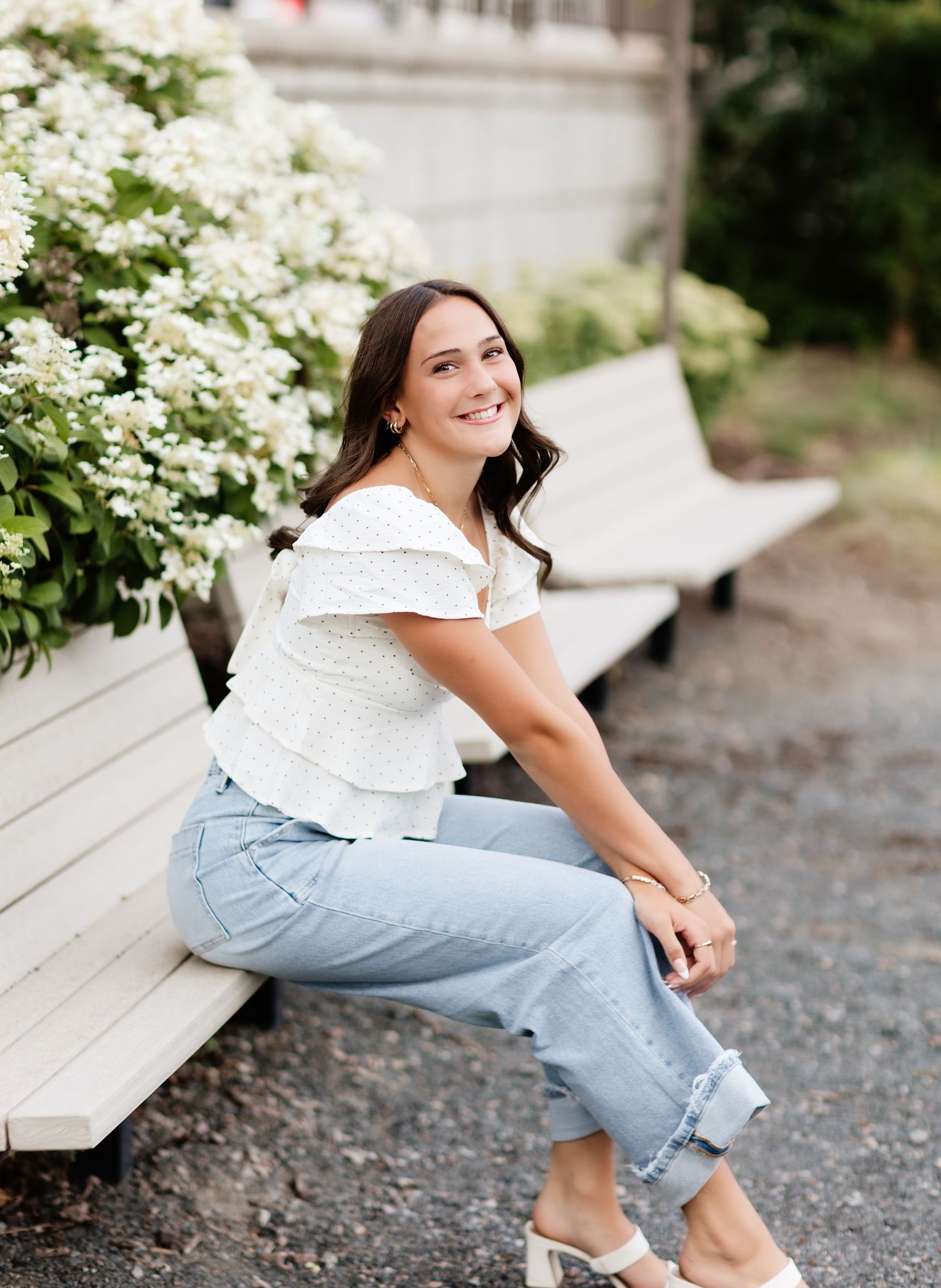 Happy young woman in white eyelet top and jeans sitting on a bench beside blooming white hydrangeas.