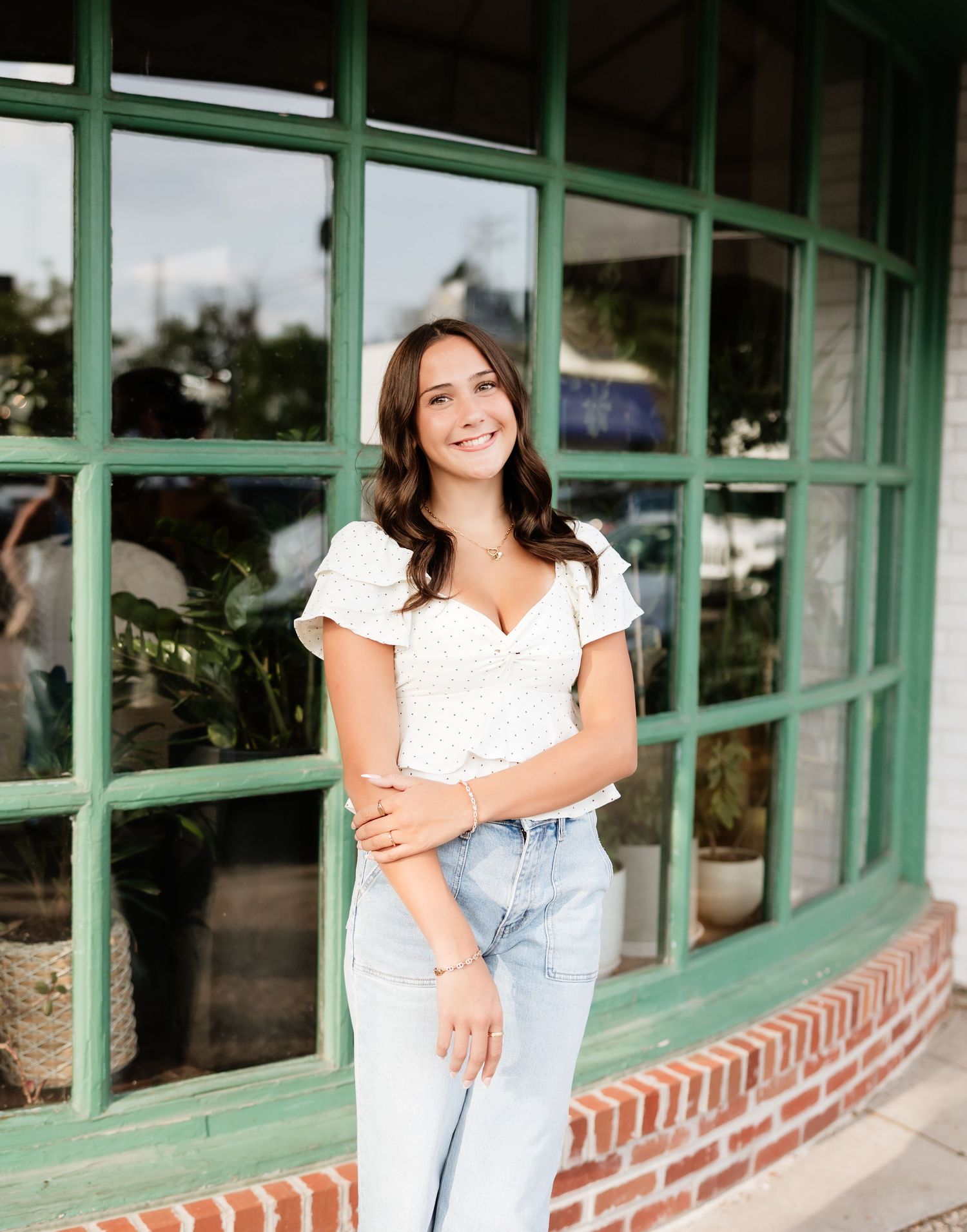 Brunette woman in white puff-sleeve top and light jeans smiles outside a green-trimmed shop window.