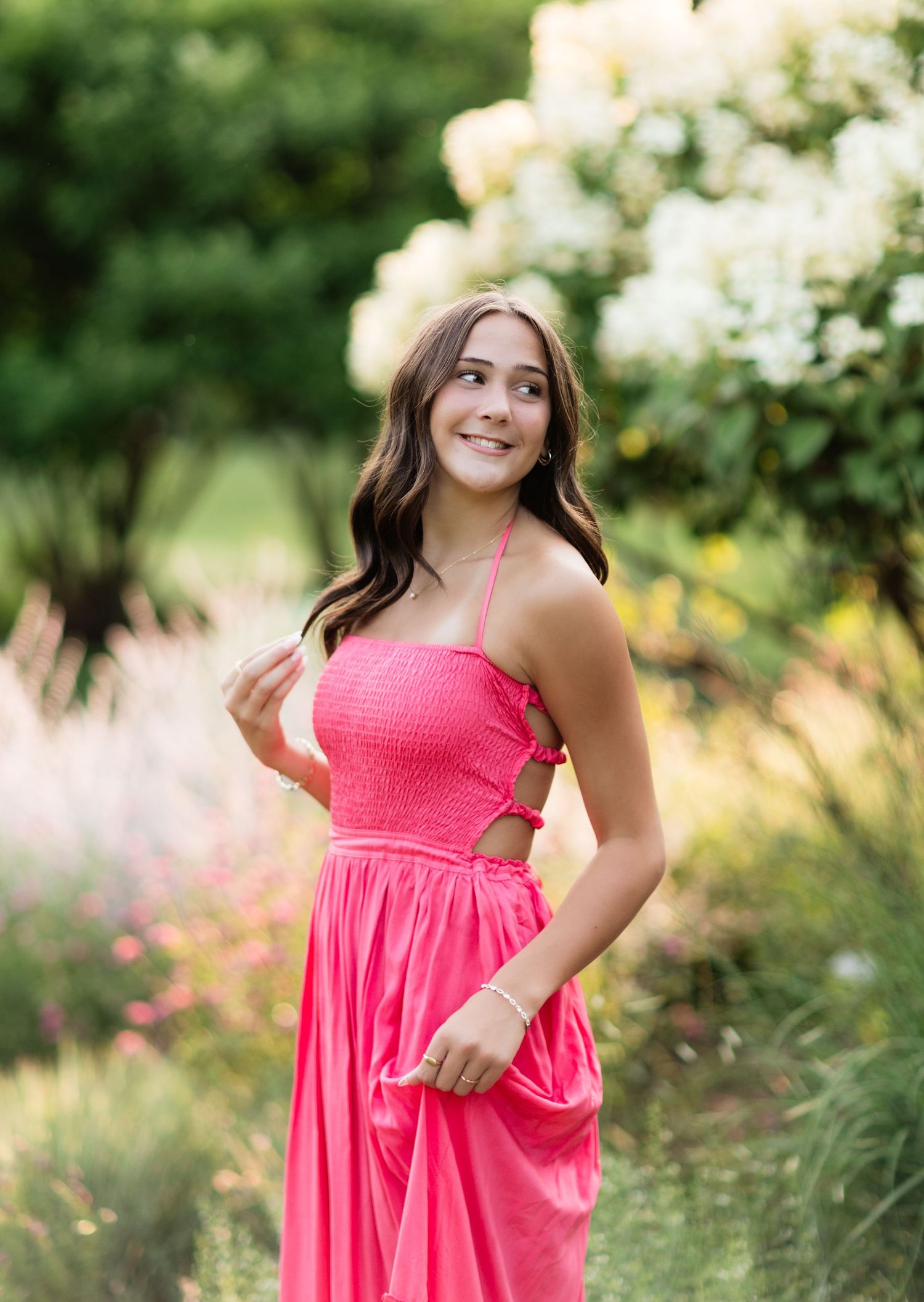 Young woman in pink smocked dress with cutout detail posing in vibrant summer flower garden.