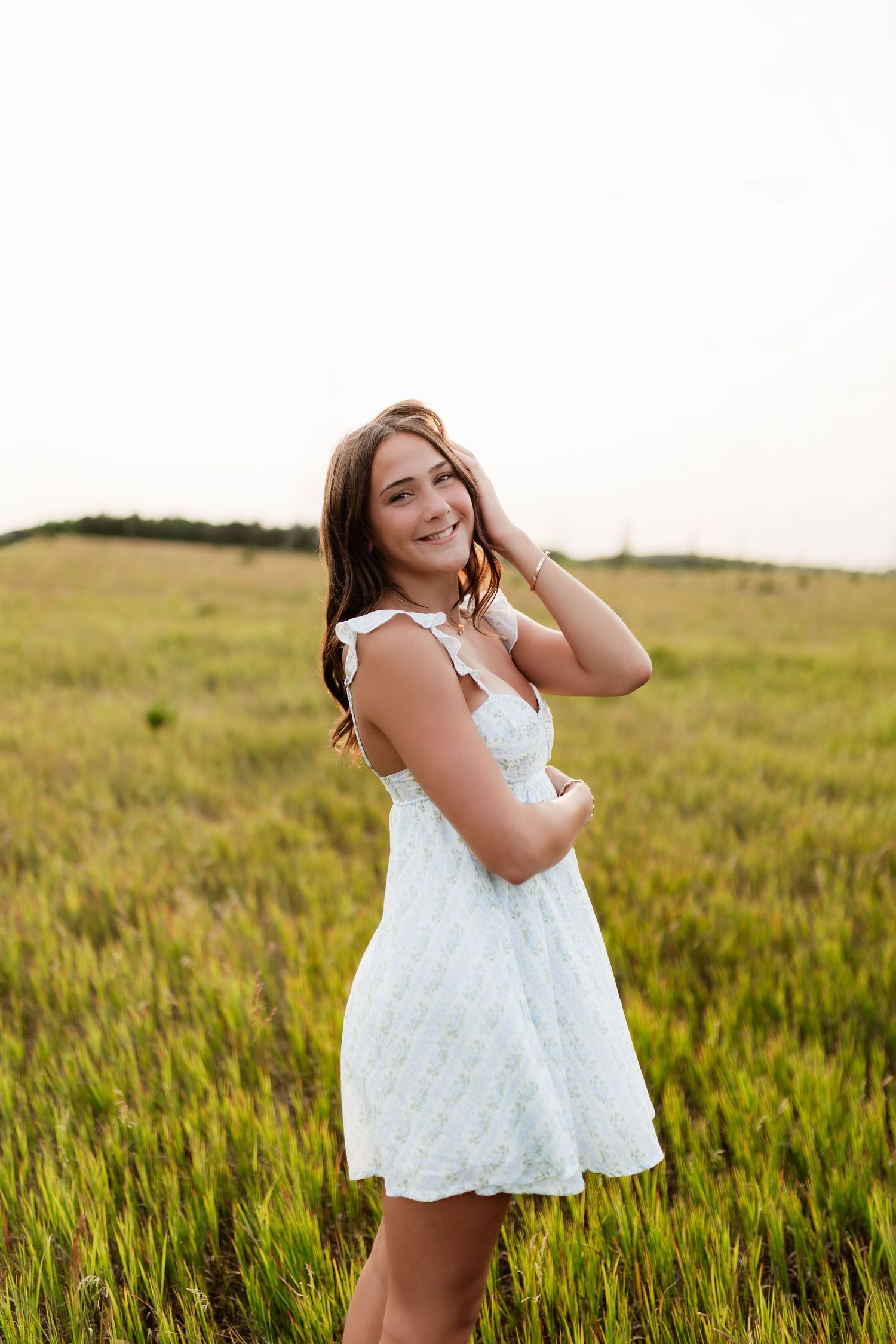 Young woman in white mini dress smiling in a lush green field at sunset.
