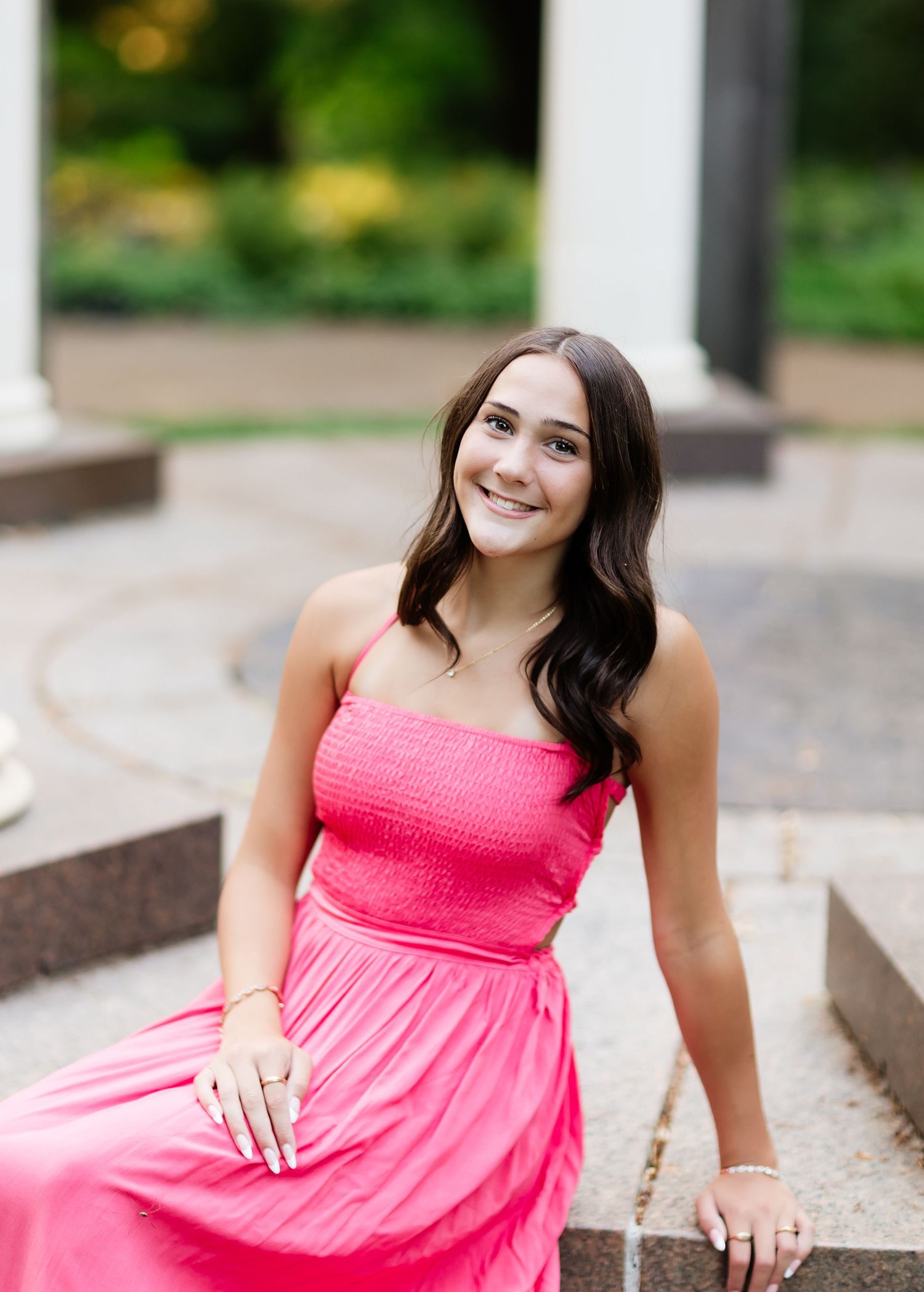 Young woman in pink maxi dress smiling outdoors on stone steps with green garden background.