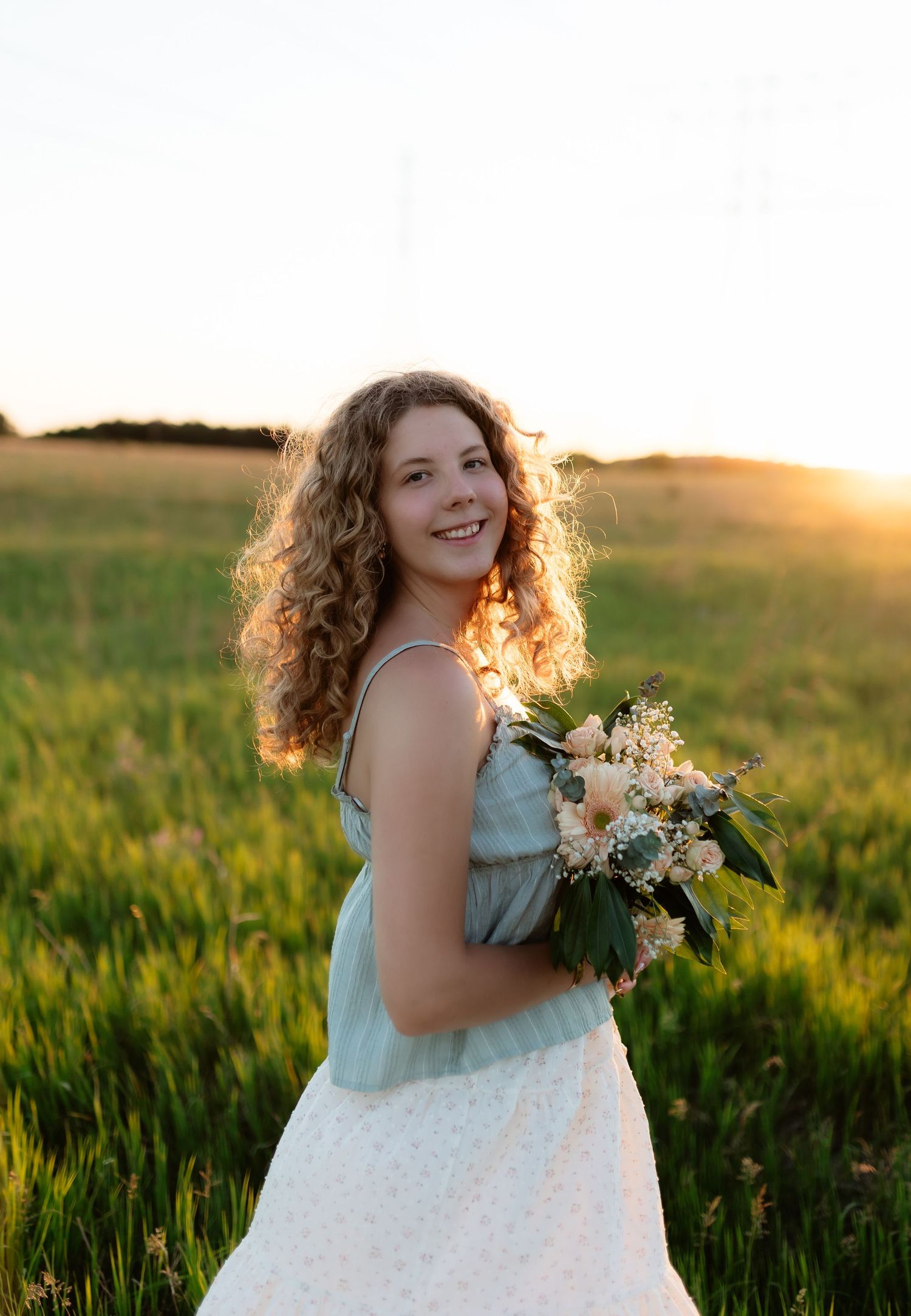 Smiling young woman with curly hair holding a bouquet of flowers in a sunlit green field.