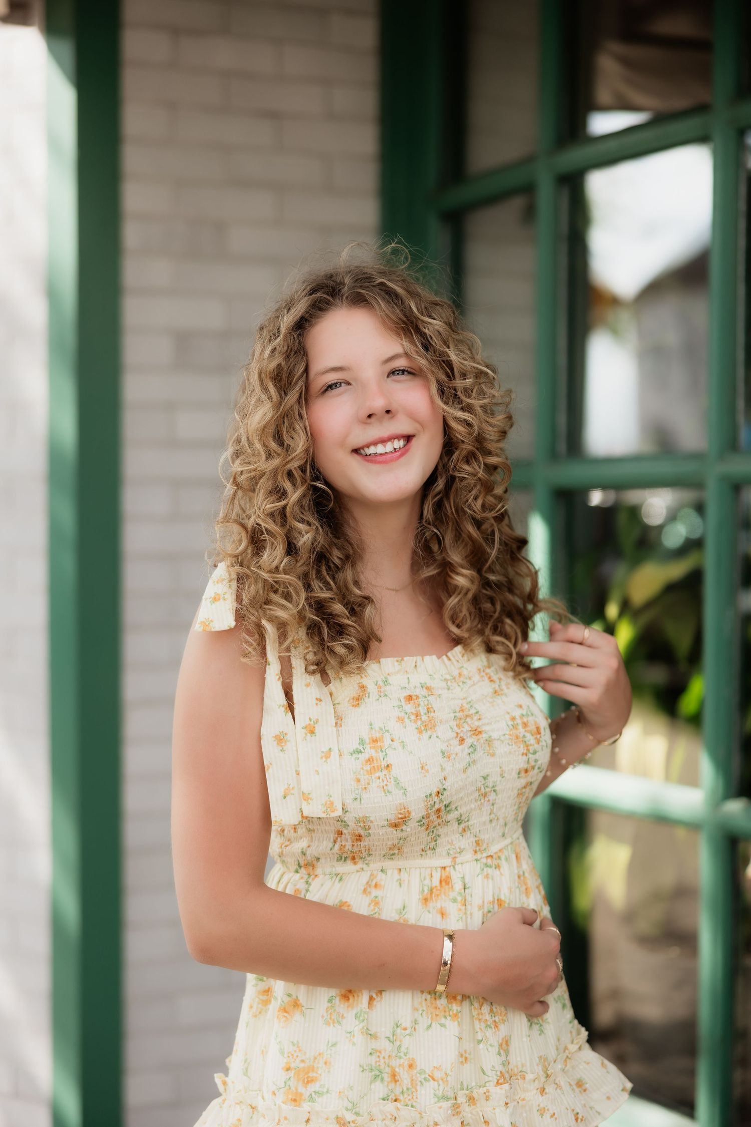 Teenage girl with curly hair smiling in yellow floral sundress standing near green-framed window.