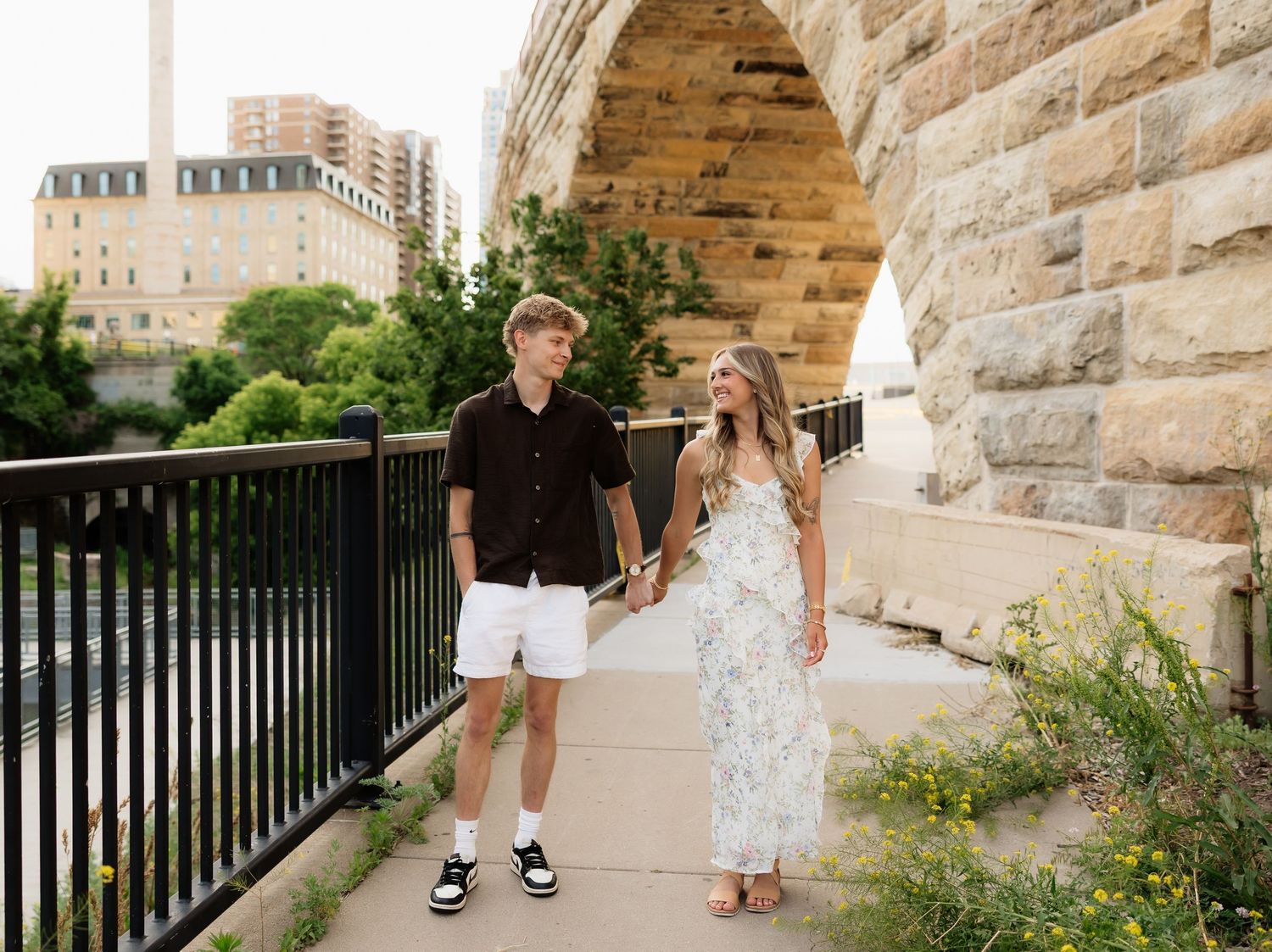 A couple holding hands and walking along a stone arch bridge pathway with city buildings visible in the background.