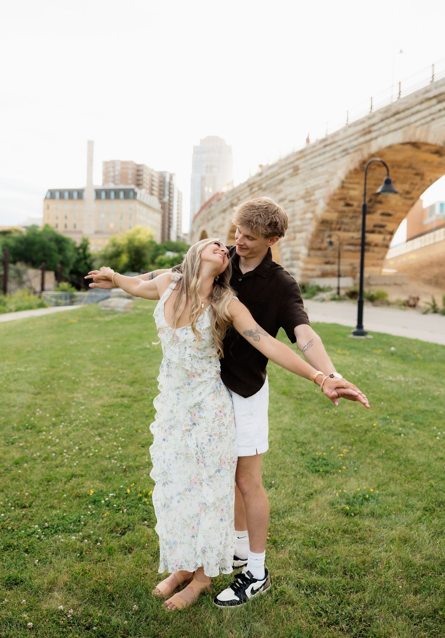 Happy couple dancing on green grass with historic stone arch bridge in background, woman in floral dress, man in black polo.