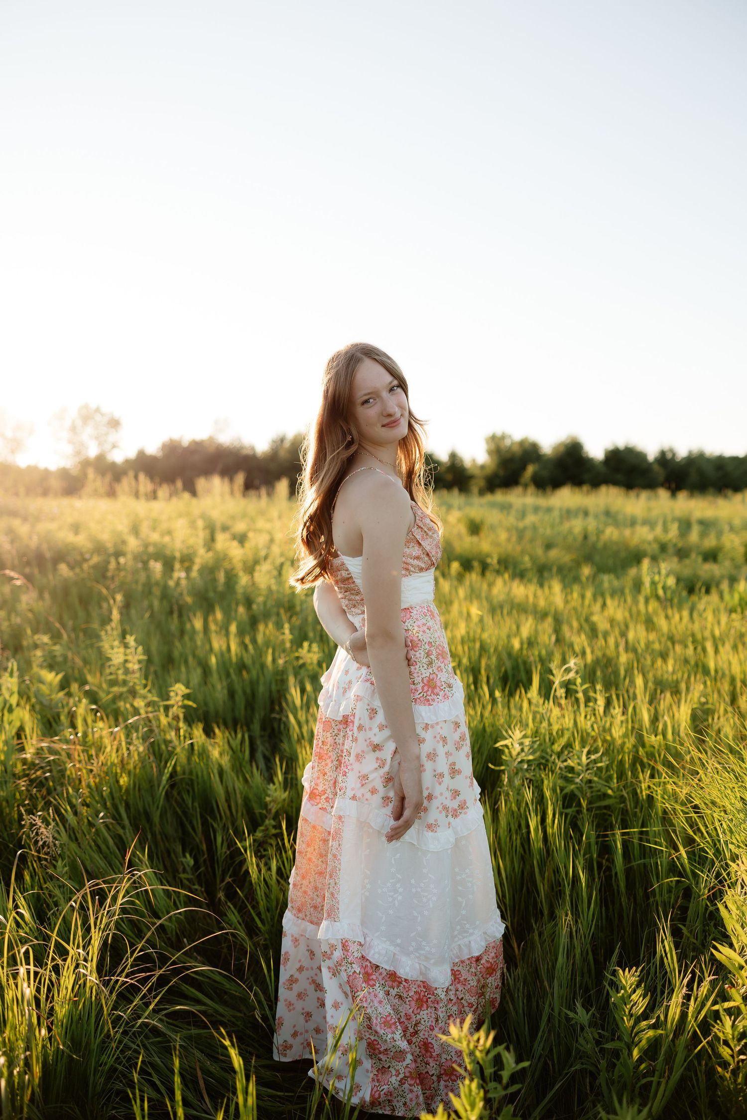 Young woman in floral maxi dress standing in green field at golden sunset hour.