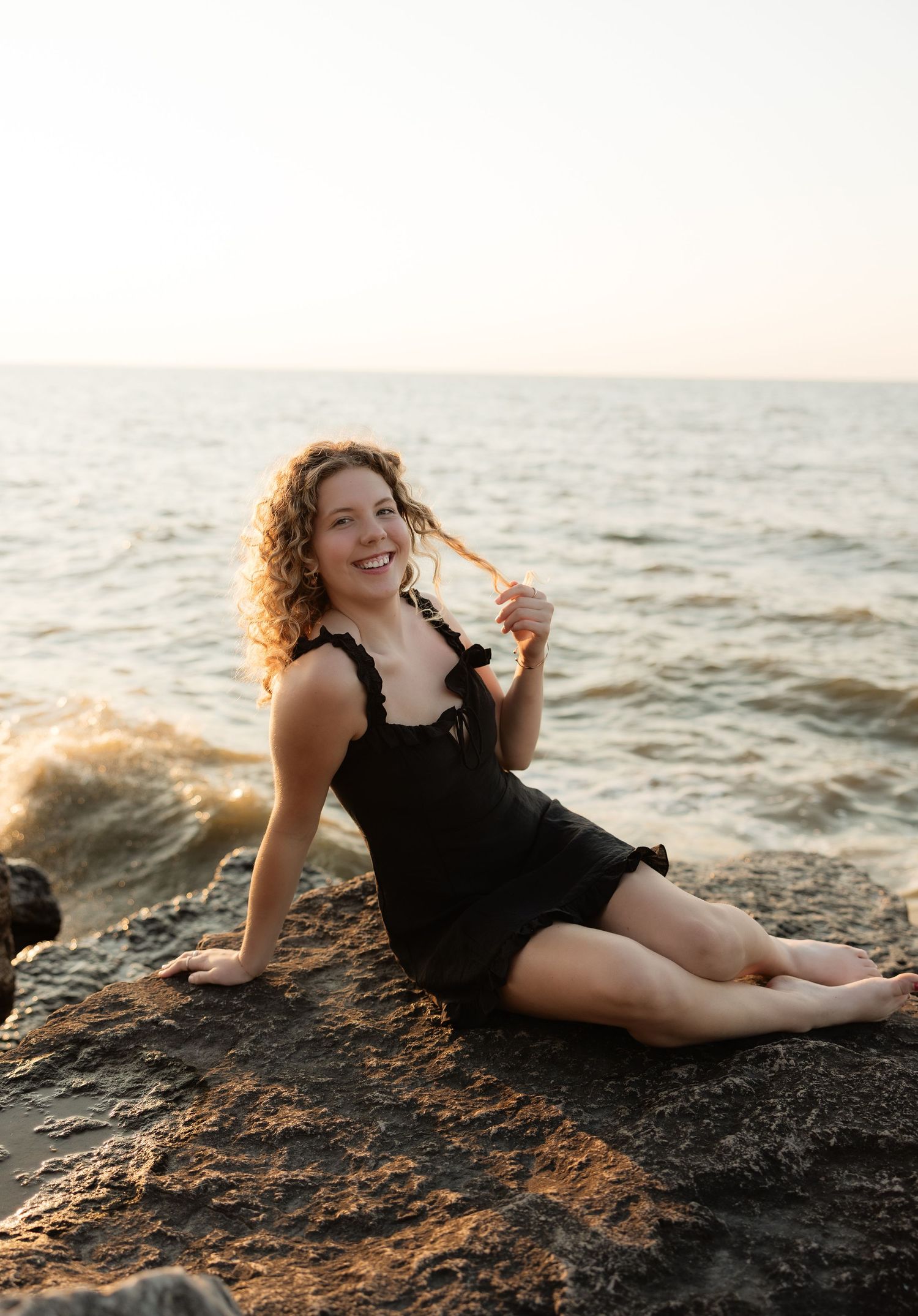 Woman in black dress sitting on rocks by the ocean at sunset, smiling and twirling her curly hair.