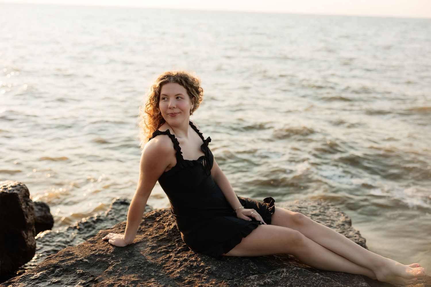 Woman in black dress sitting on rocks by the ocean at sunset, smiling peacefully at the camera.