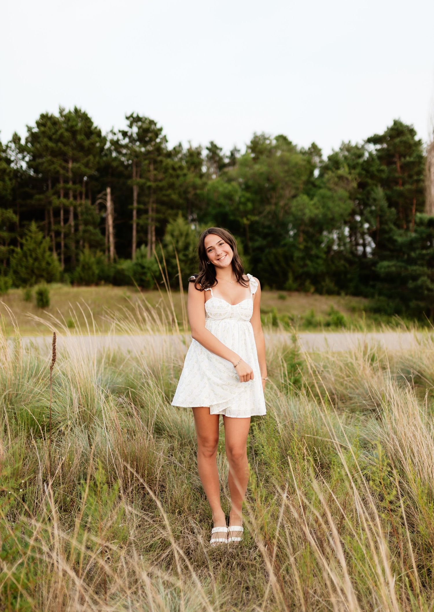 Brunette woman in white dress standing in tall grass with trees behind.