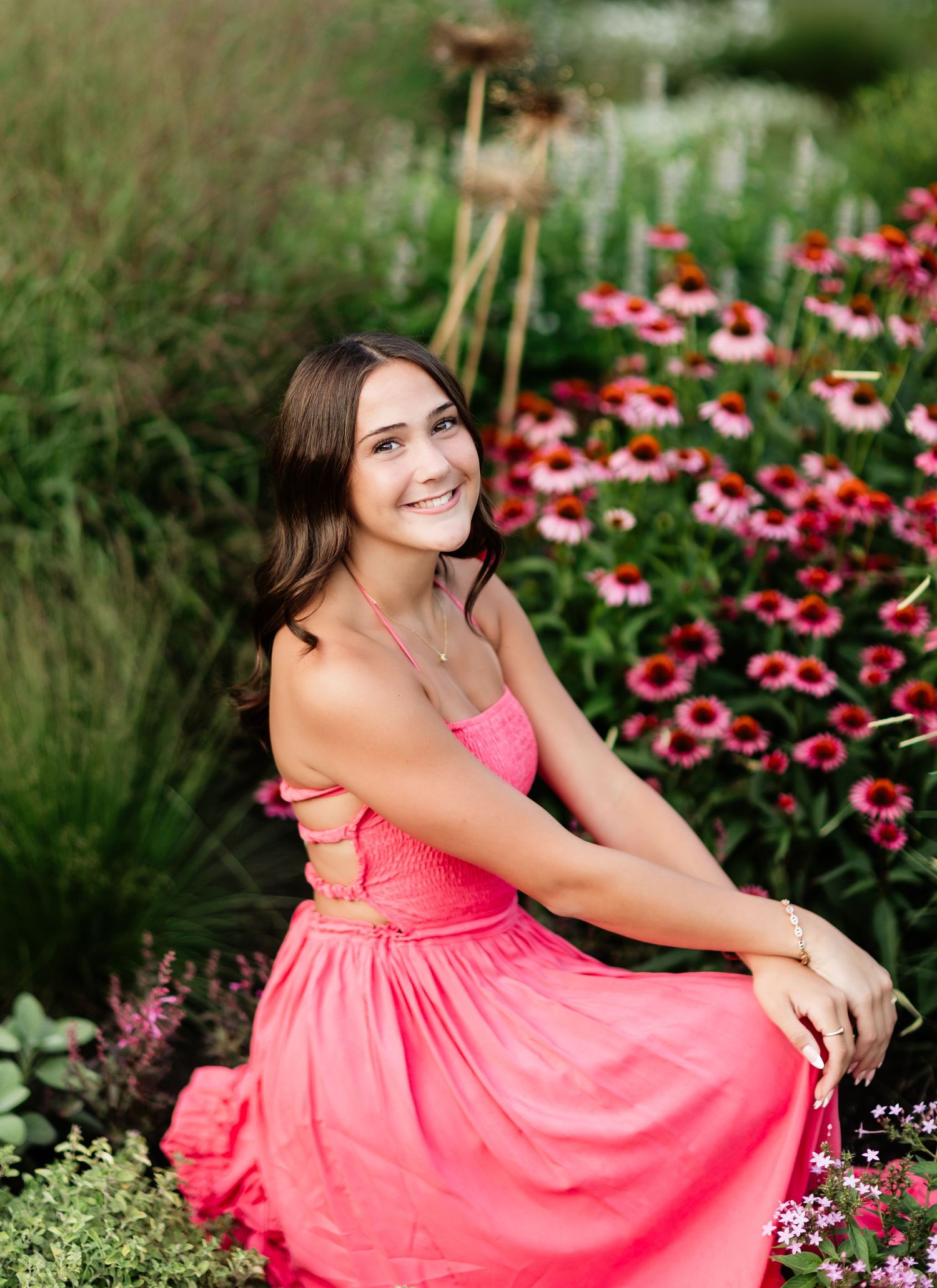 Smiling brunette in pink dress sitting among colorful coneflowers in garden.