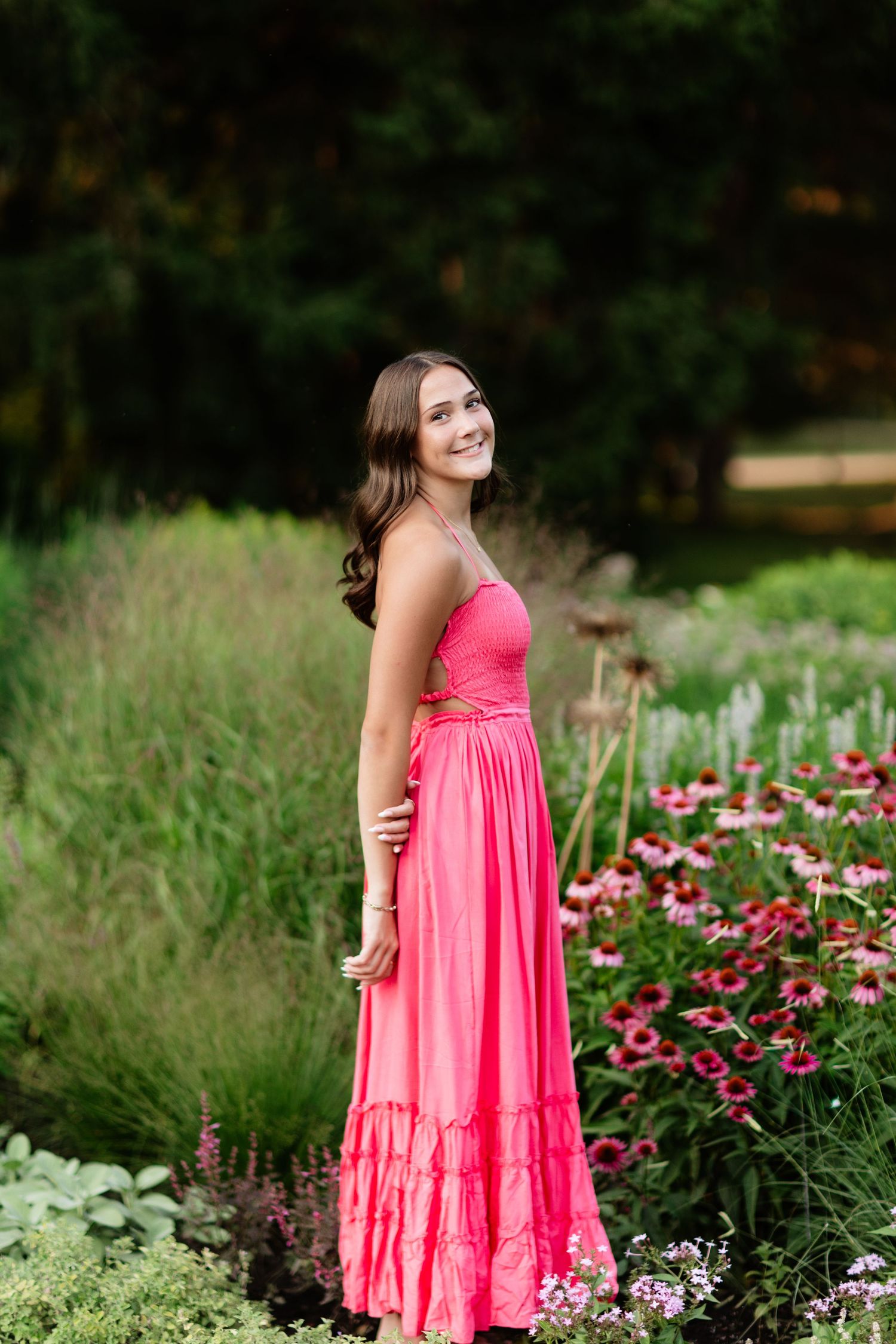 Smiling brunette in pink maxi dress standing beside colorful garden flowers in soft light.