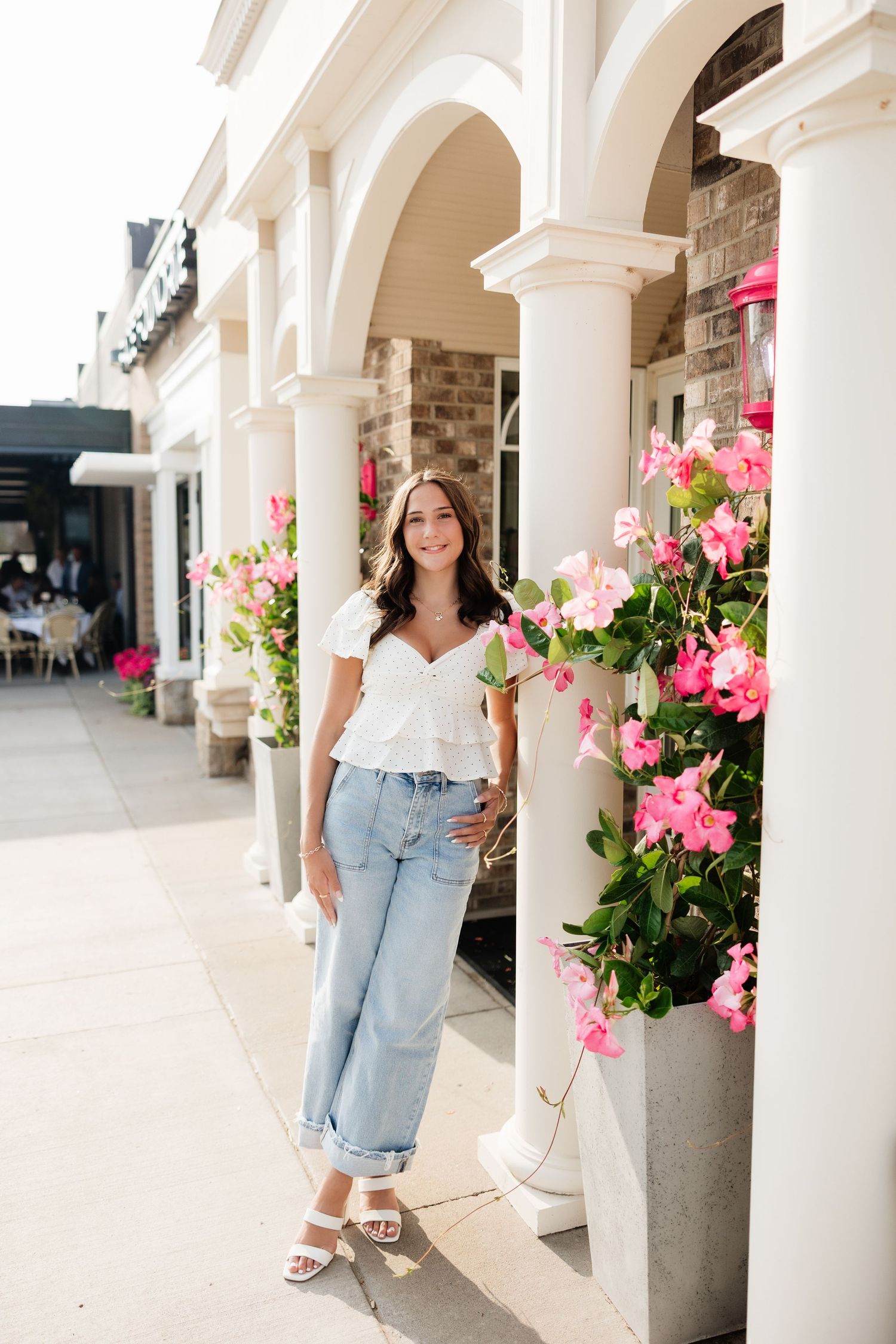 Smiling young woman in white top and jeans posing near pink flowers and white columns outdoors.