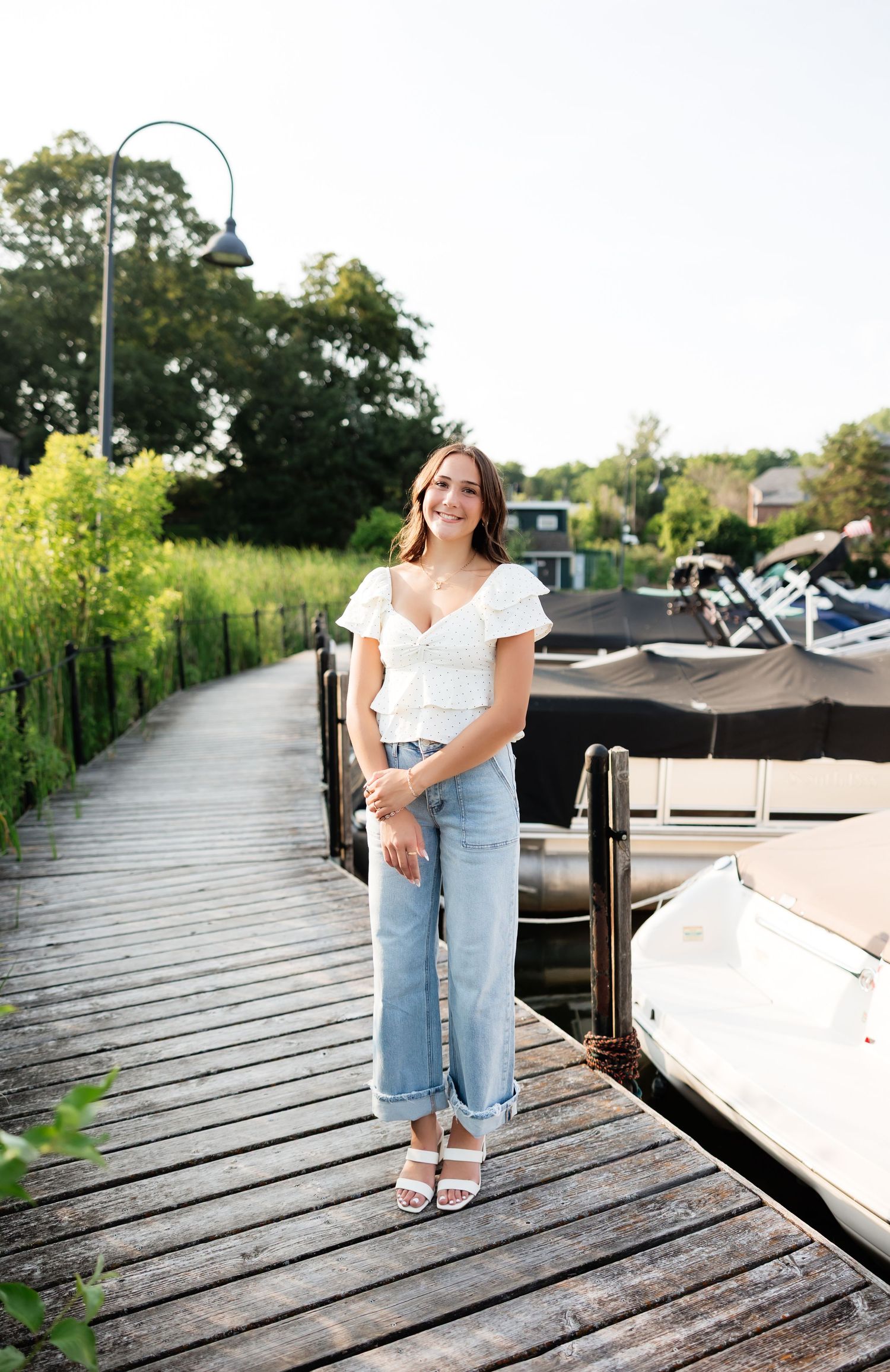 Smiling young woman in white top and jeans stands on a wooden marina dock with boats nearby.