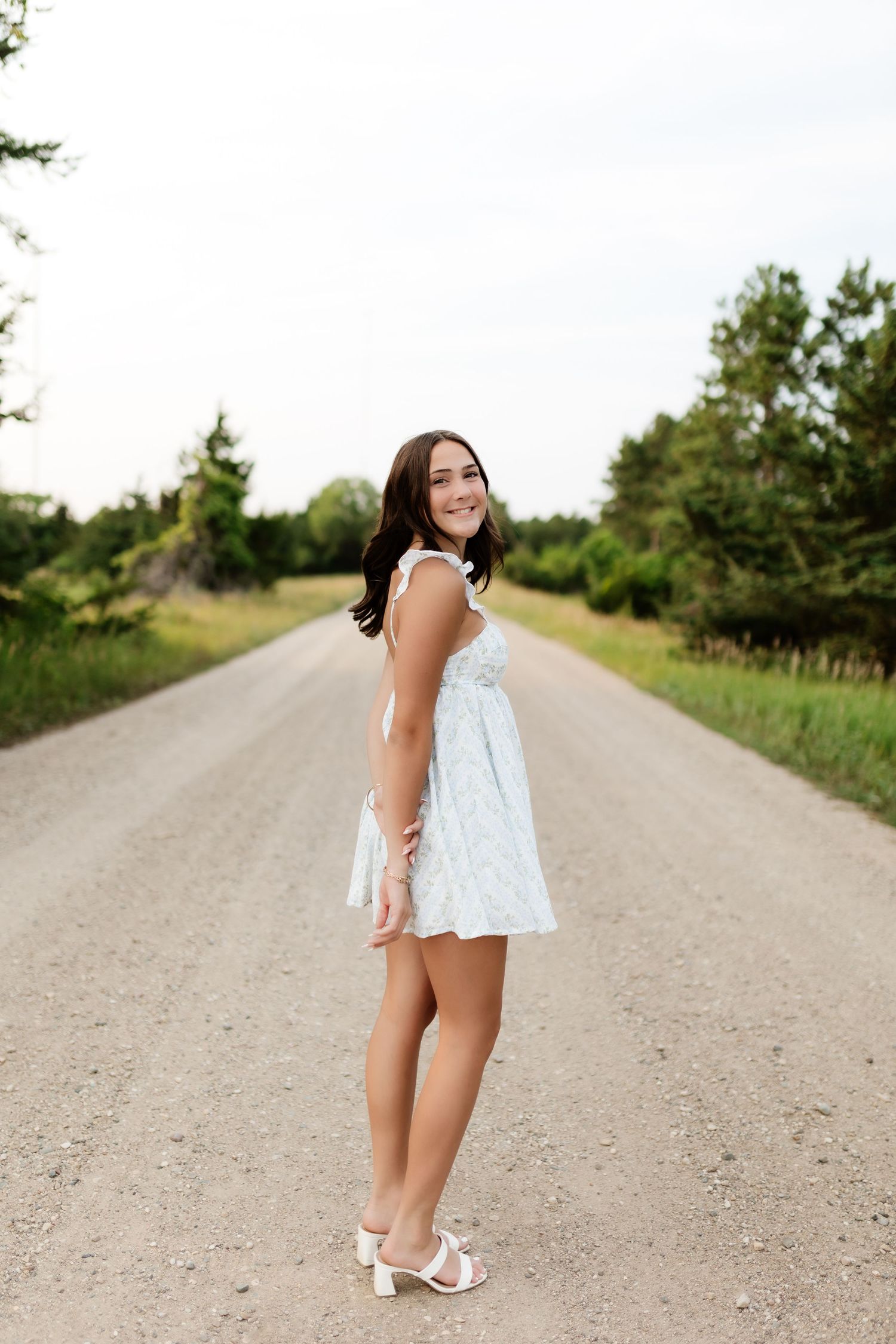 Brunette teen in white dress smiling on a dirt road lined with green trees.