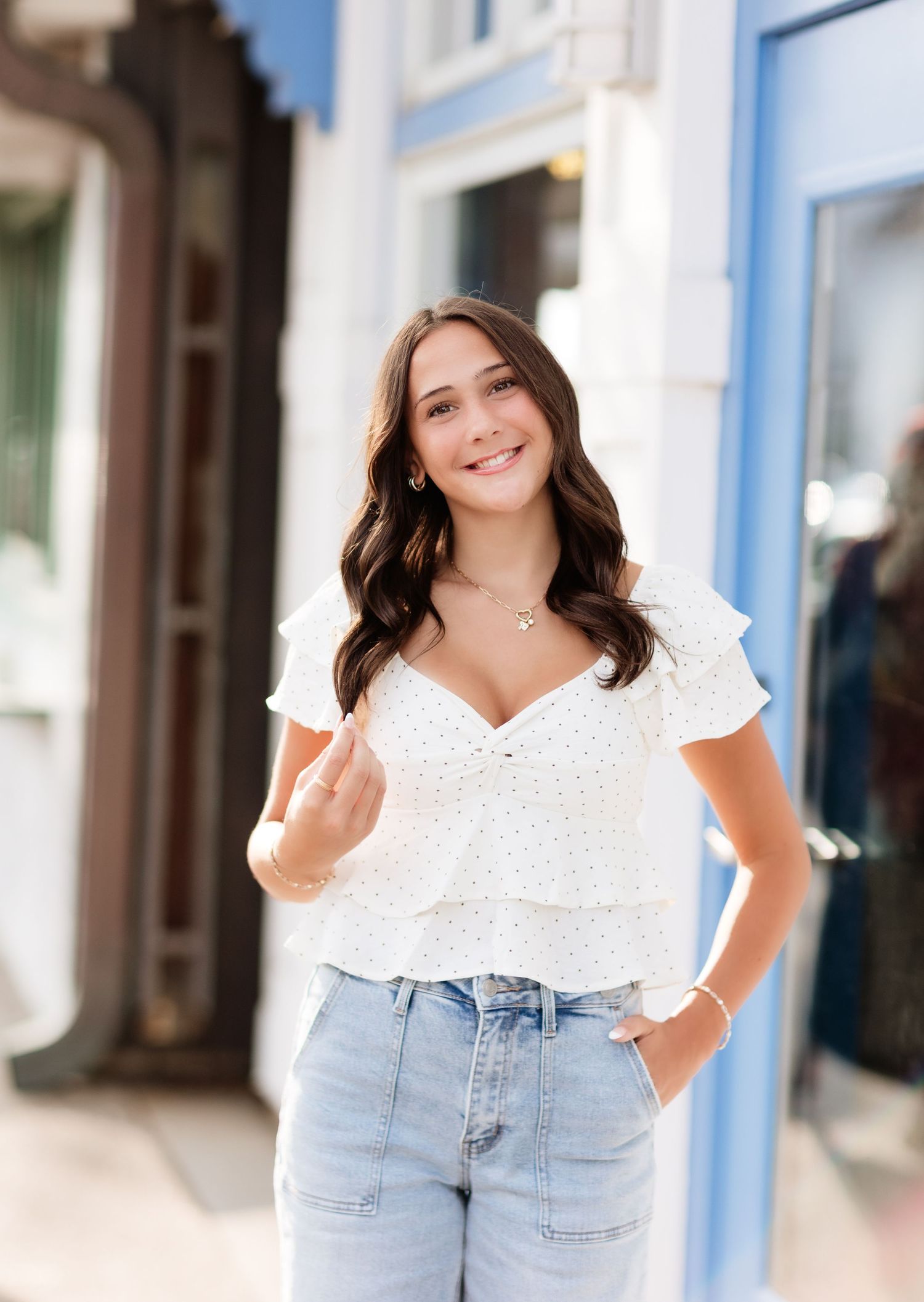 Brunette woman in white eyelet top and jeans smiling confidently outside blue storefront building.