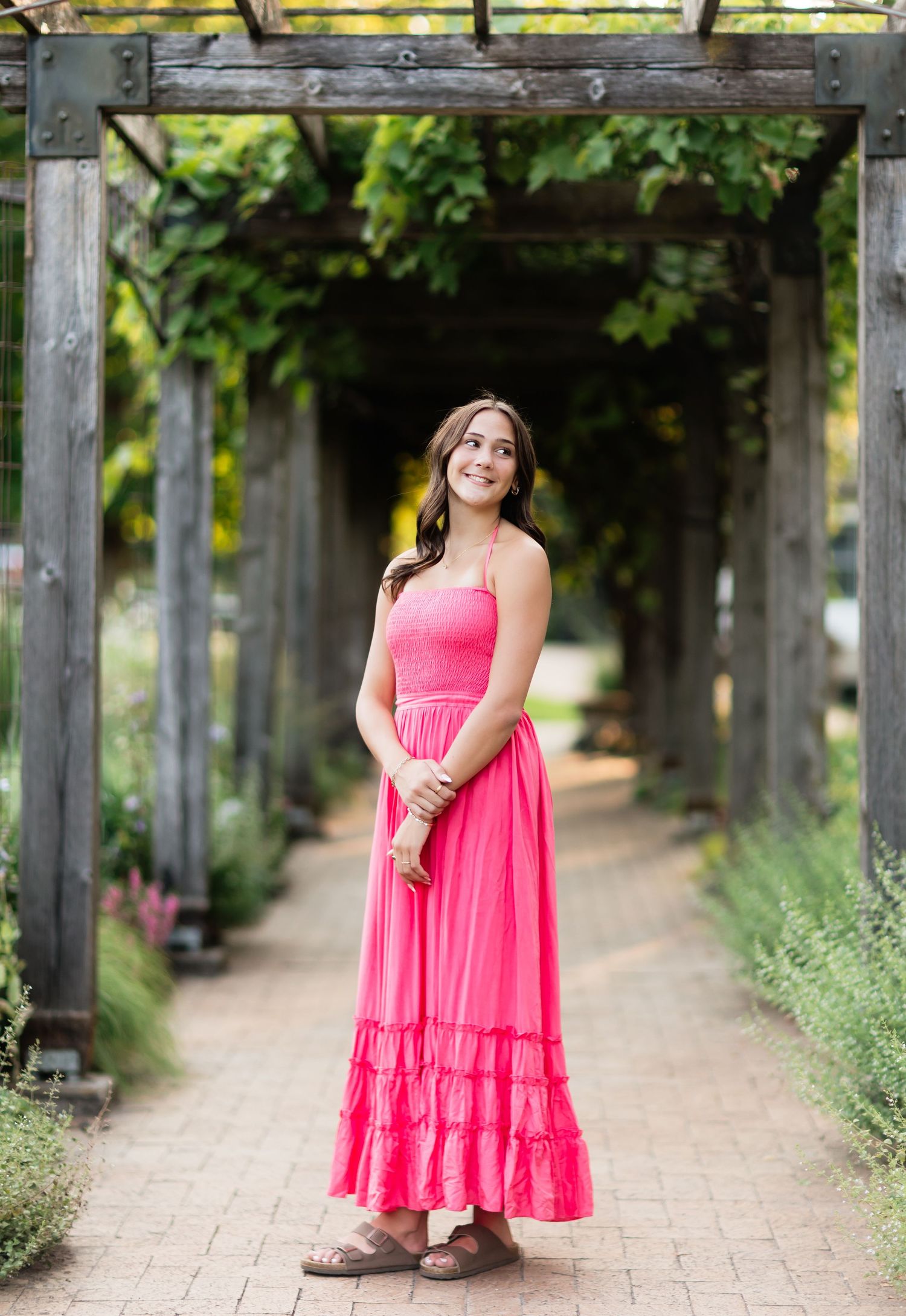Smiling young woman in hot pink maxi dress standing under a wooden pergola walkway in a garden.