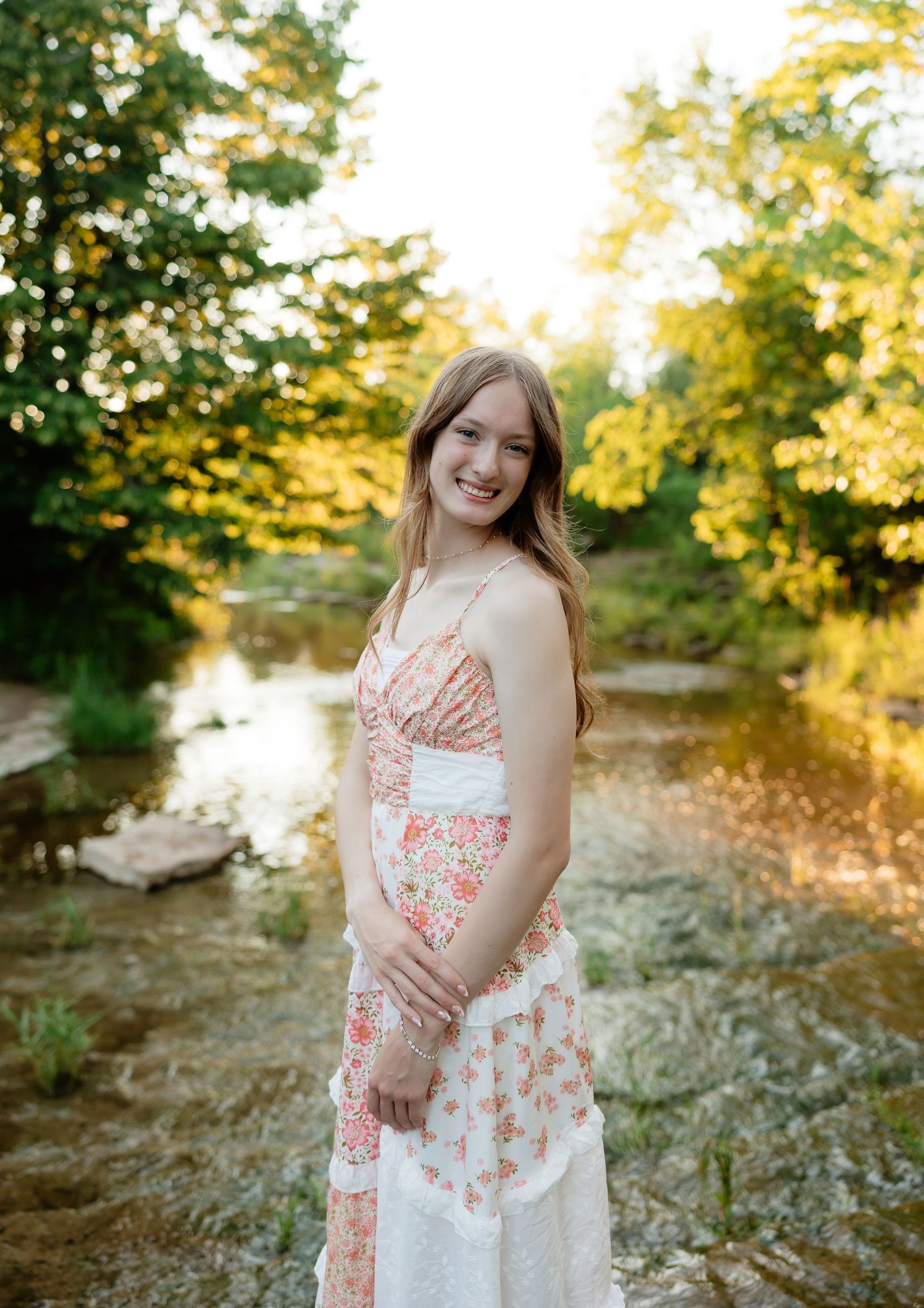 Young woman in floral dress smiling by a sunlit creek surrounded by trees.