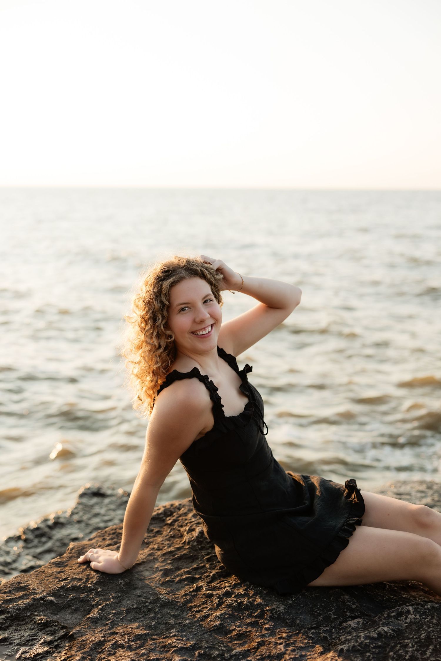 A smiling woman with curly hair in a black dress sits on rocks by the ocean at sunset.