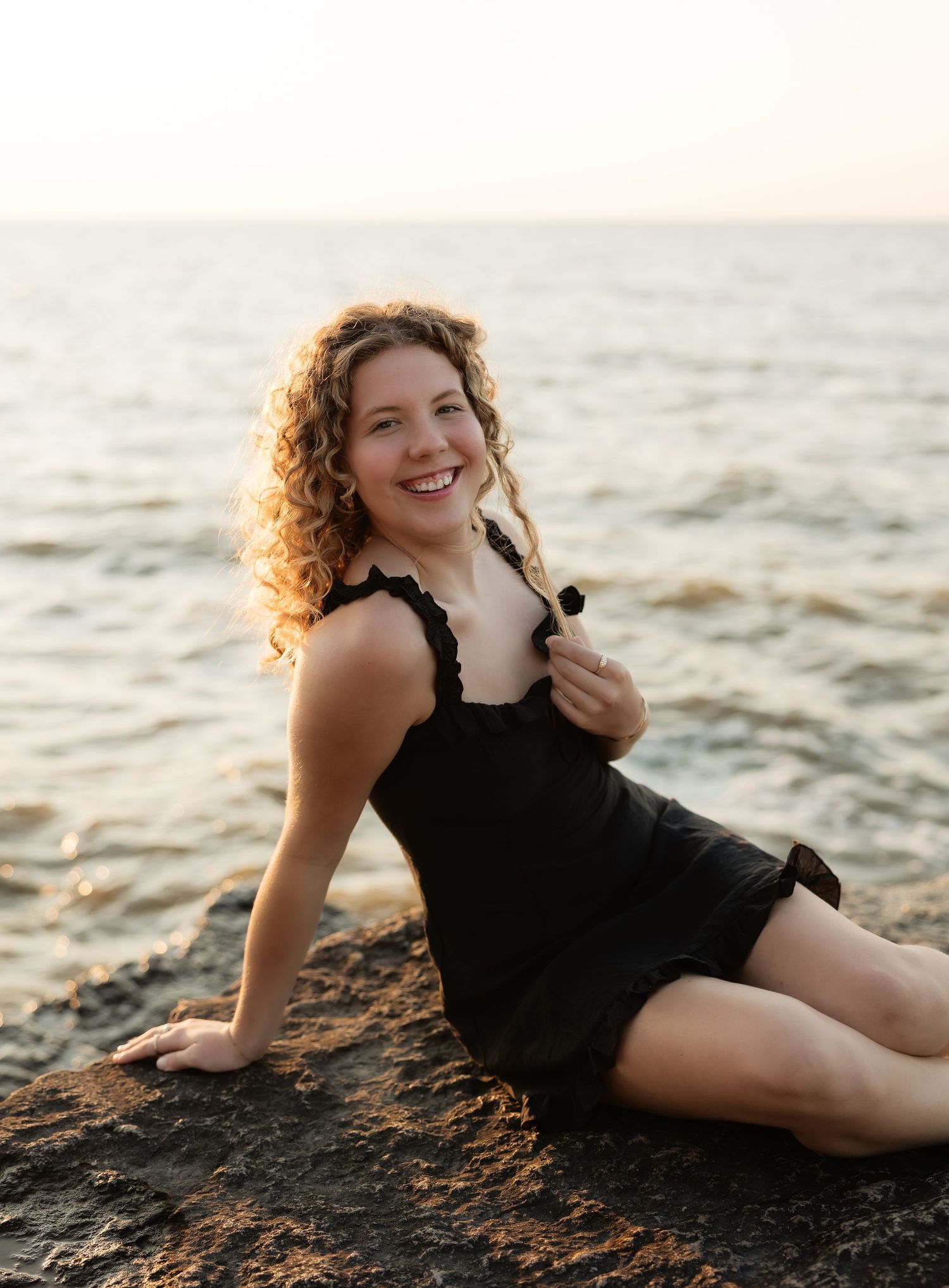 Smiling young woman with curly hair in black dress sitting on rocks by the ocean at sunset.