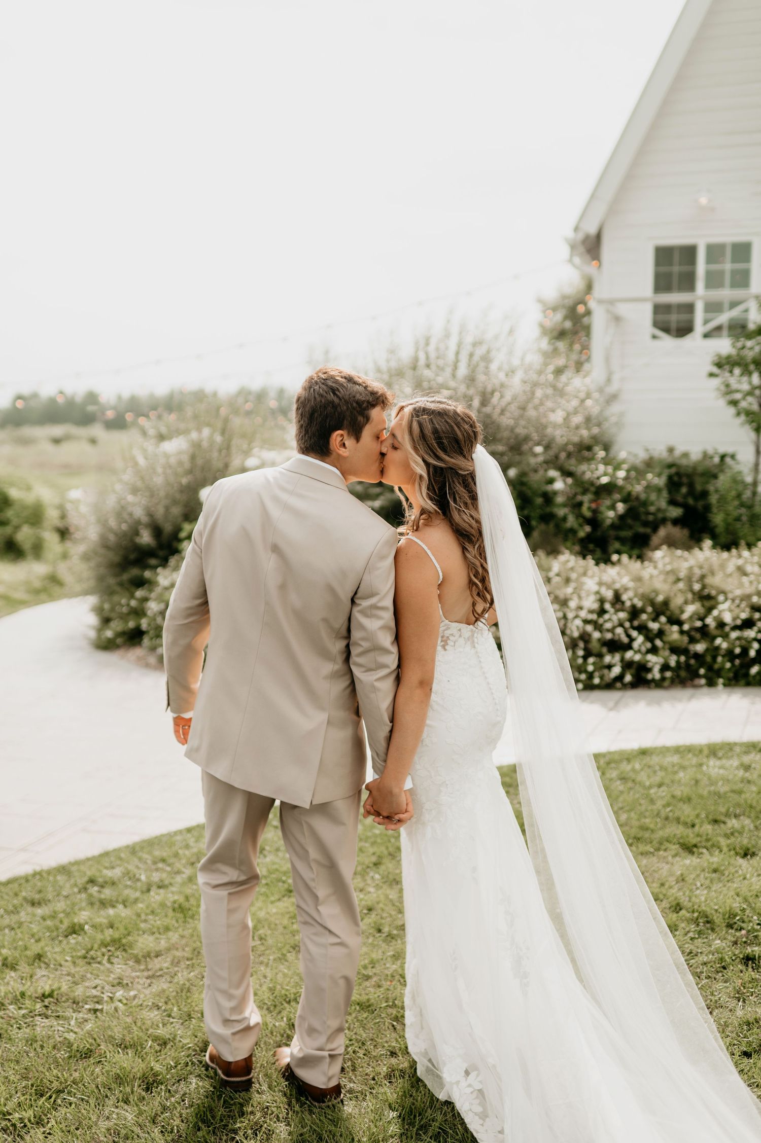 Bride and groom holding hands and kissing near a white chapel surrounded by lush greenery.