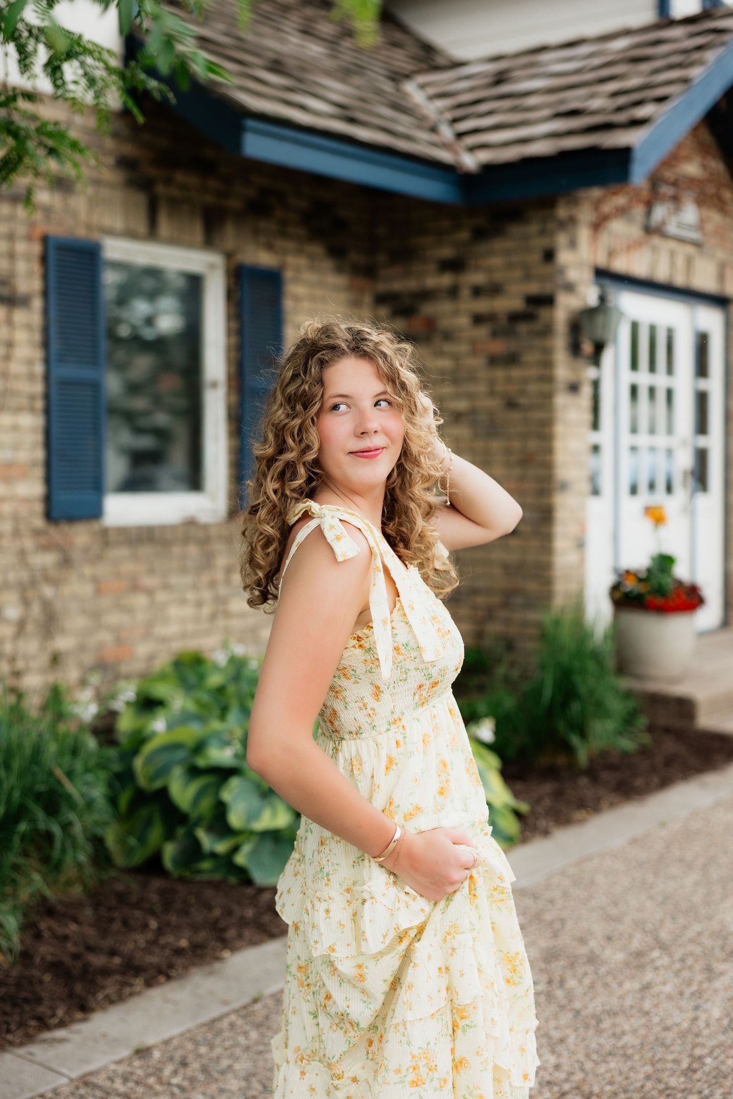 Young woman with curly hair in a floral yellow sundress posing outdoors near a brick building.