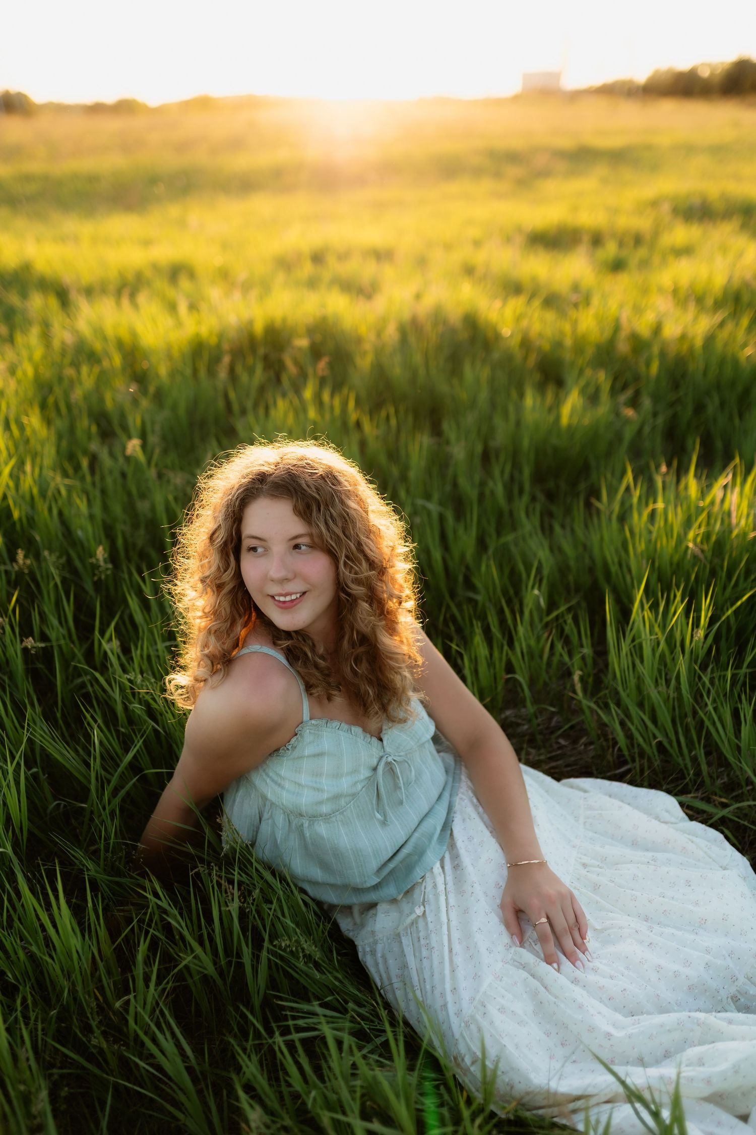 Smiling young woman in light blue dress sitting in tall green grass during a golden sunset.