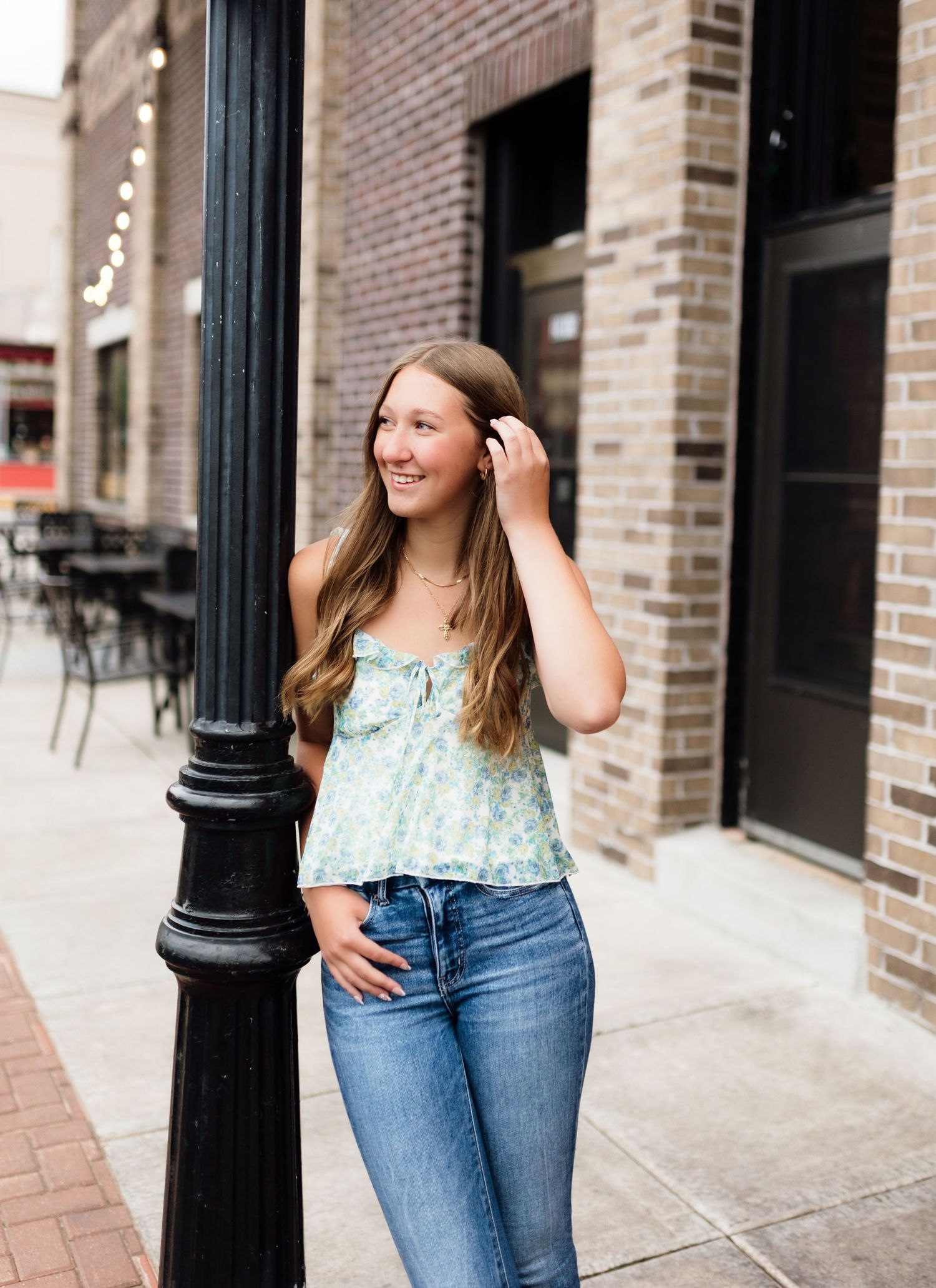 Smiling young woman in floral top and jeans leaning against a black lamppost on a brick urban sidewalk.