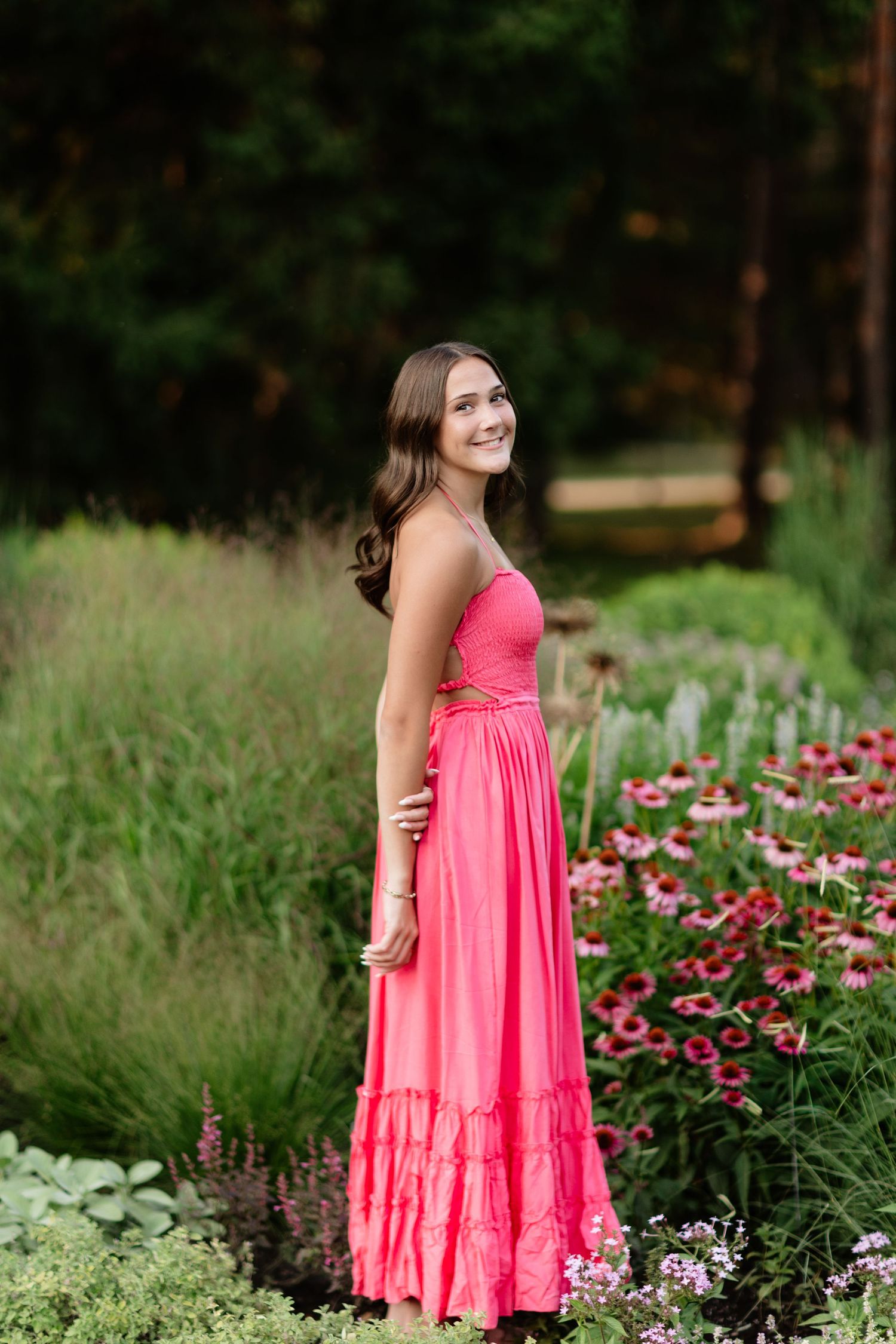 Brunette woman in coral strapless maxi dress standing among colorful garden flowers, smiling.