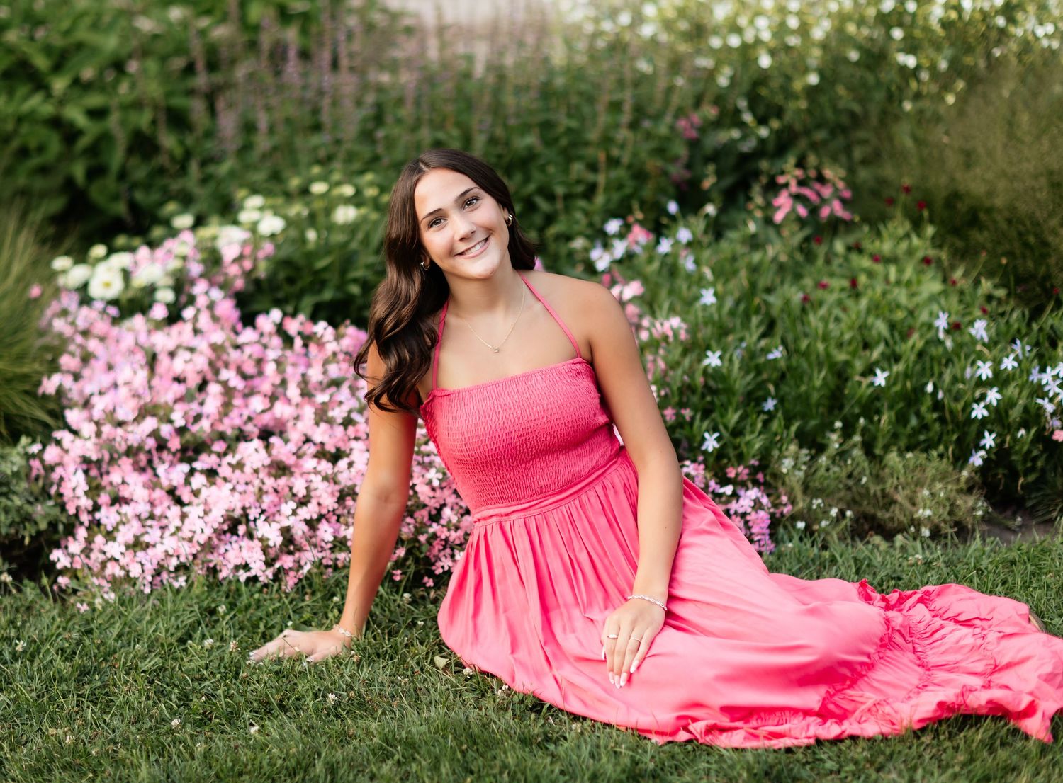 Young woman in hot pink maxi dress sitting gracefully on grass surrounded by blooming pink flowers.