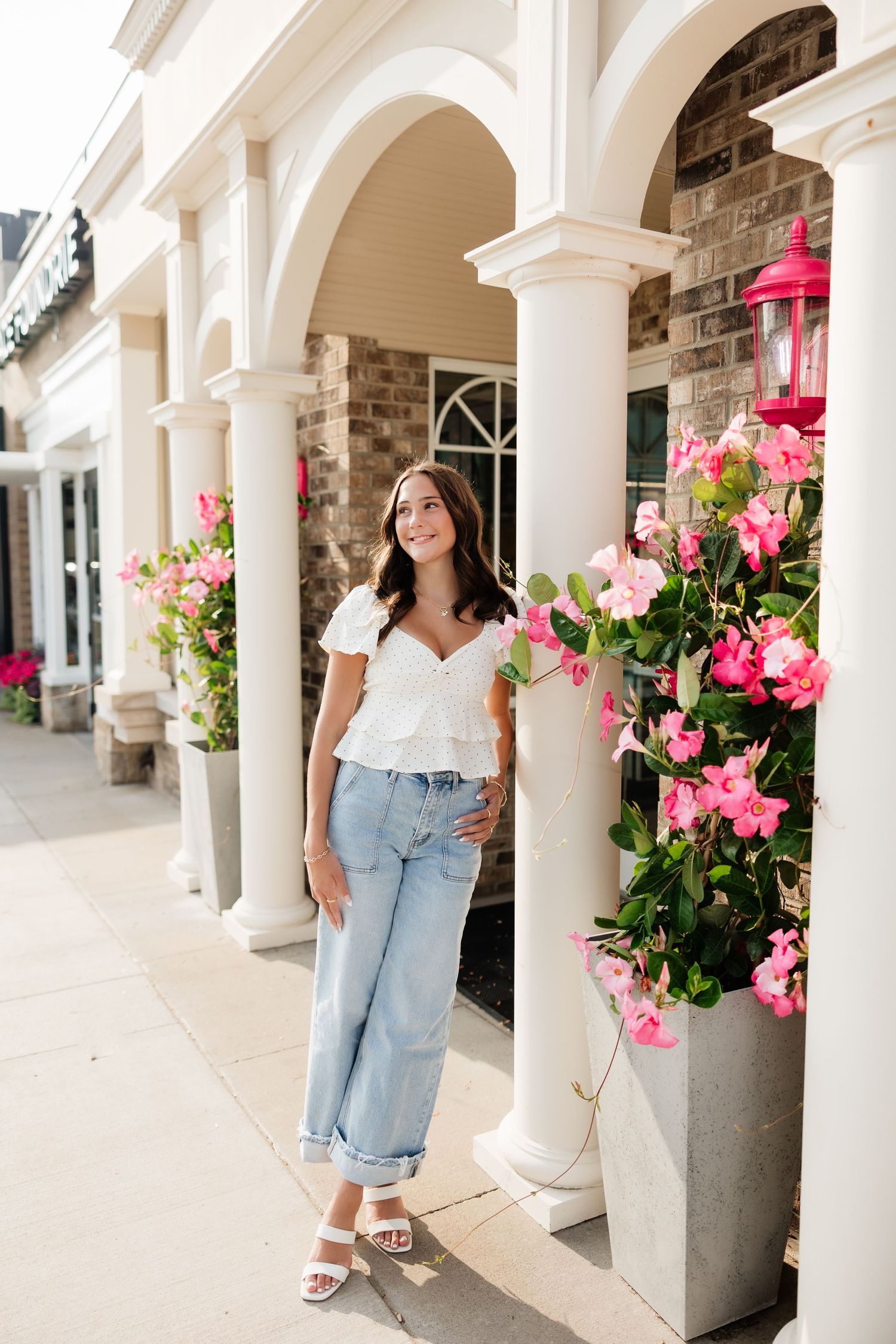 Young woman in white top and jeans poses by pink flowers and white columns at a charming outdoor shopping area.