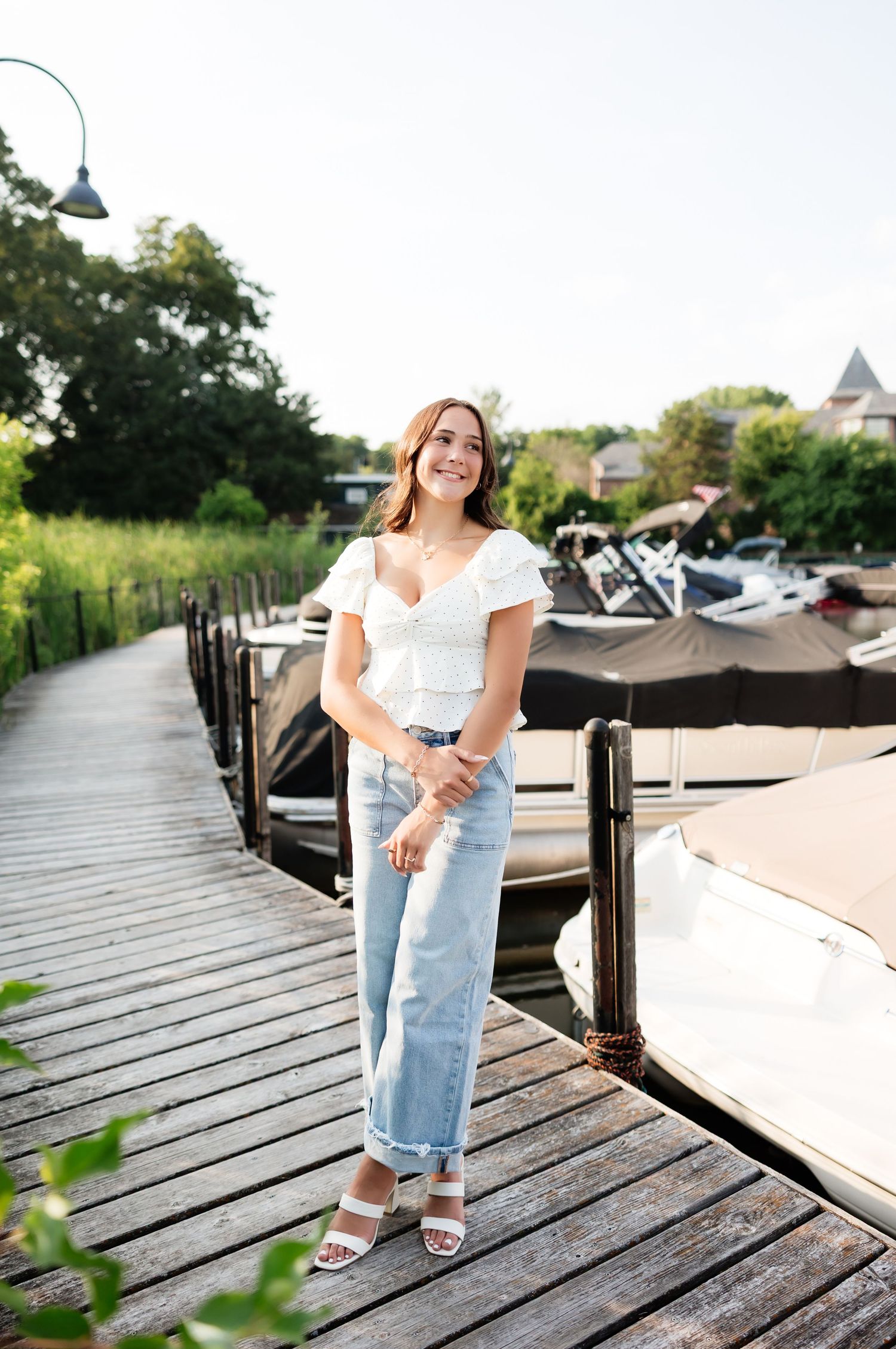 Young woman in white top and light jeans posing on a sunny wooden dock with boats nearby.