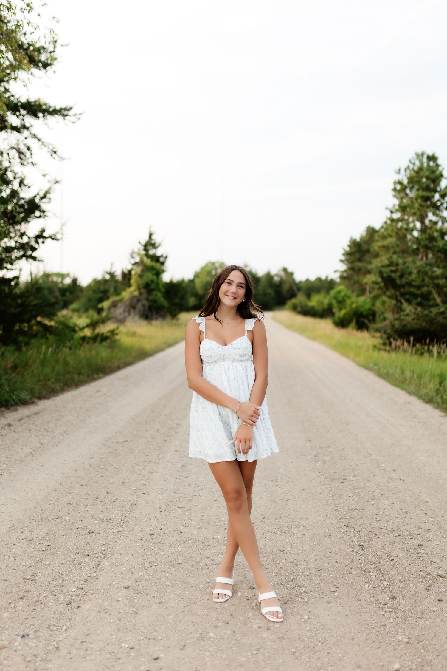 Young woman in white sundress smiling on a straight tree-lined gravel road outdoors.