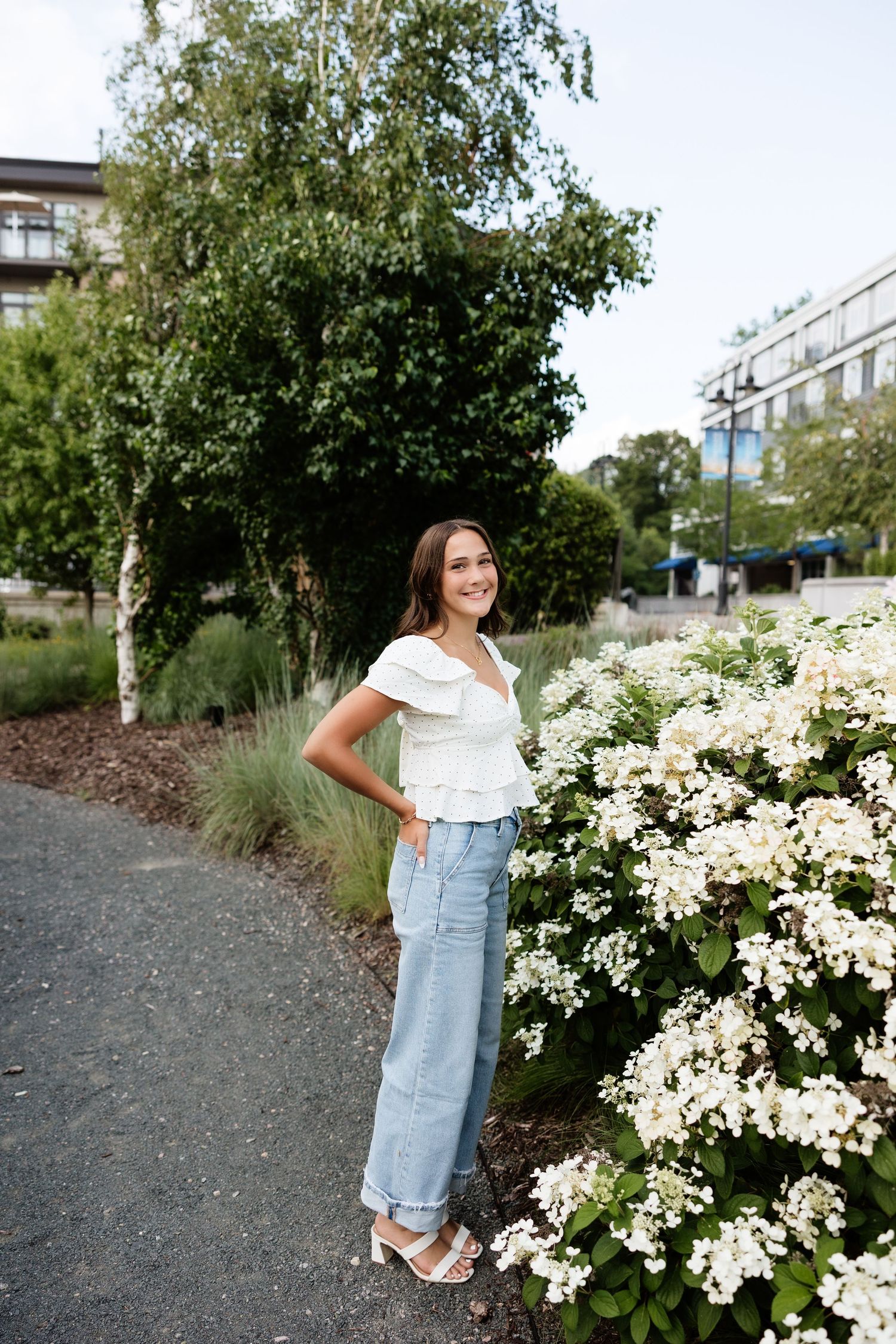 Young woman in white ruffled top and blue jeans smiling beside white hydrangea bushes in a lush green park.
