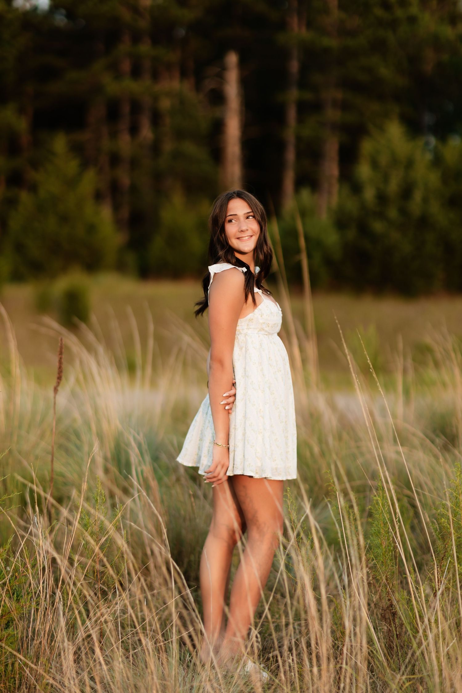 Brunette woman in white sundress standing in tall grass field at golden hour sunset.