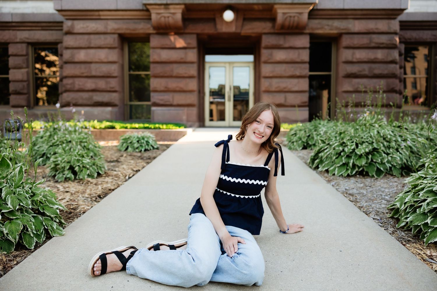 A young woman in a black top and jeans sits smiling on a pathway outside a brick building surrounded by greenery.