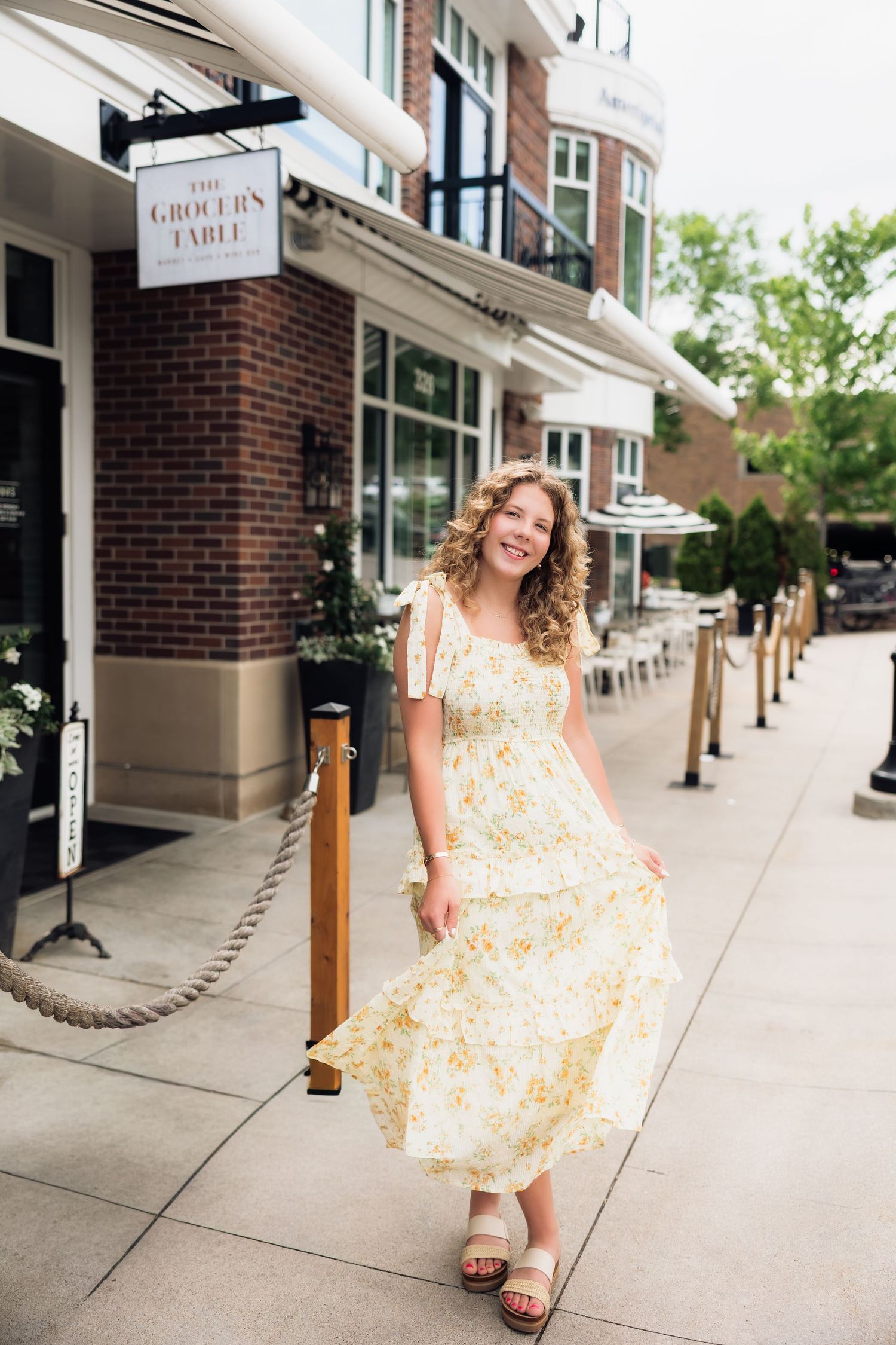 Smiling young woman in yellow floral maxi dress posing outside The Grocer's Table restaurant.