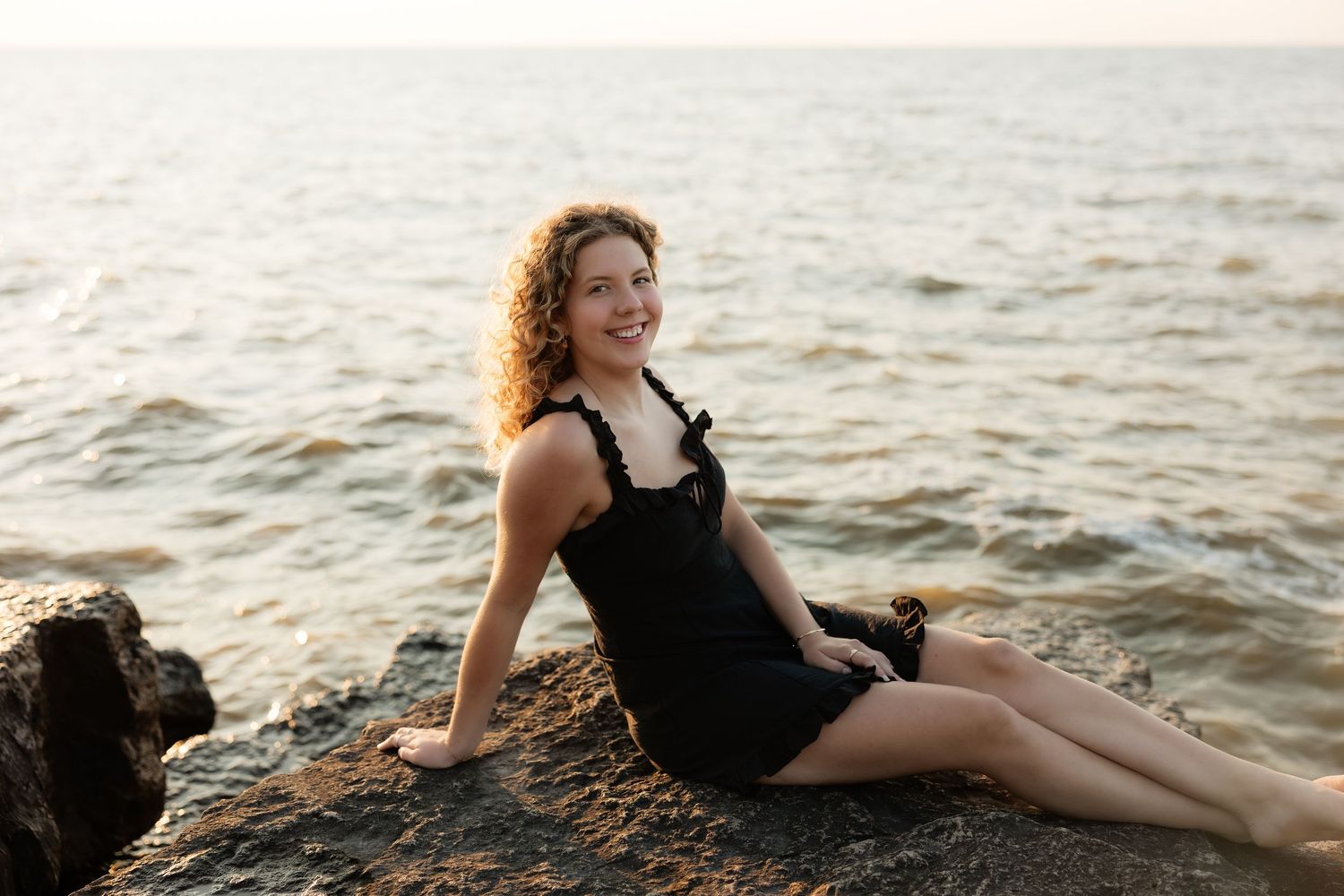 Smiling woman with curly hair in black dress sitting on rocky shore by calm ocean at sunset.