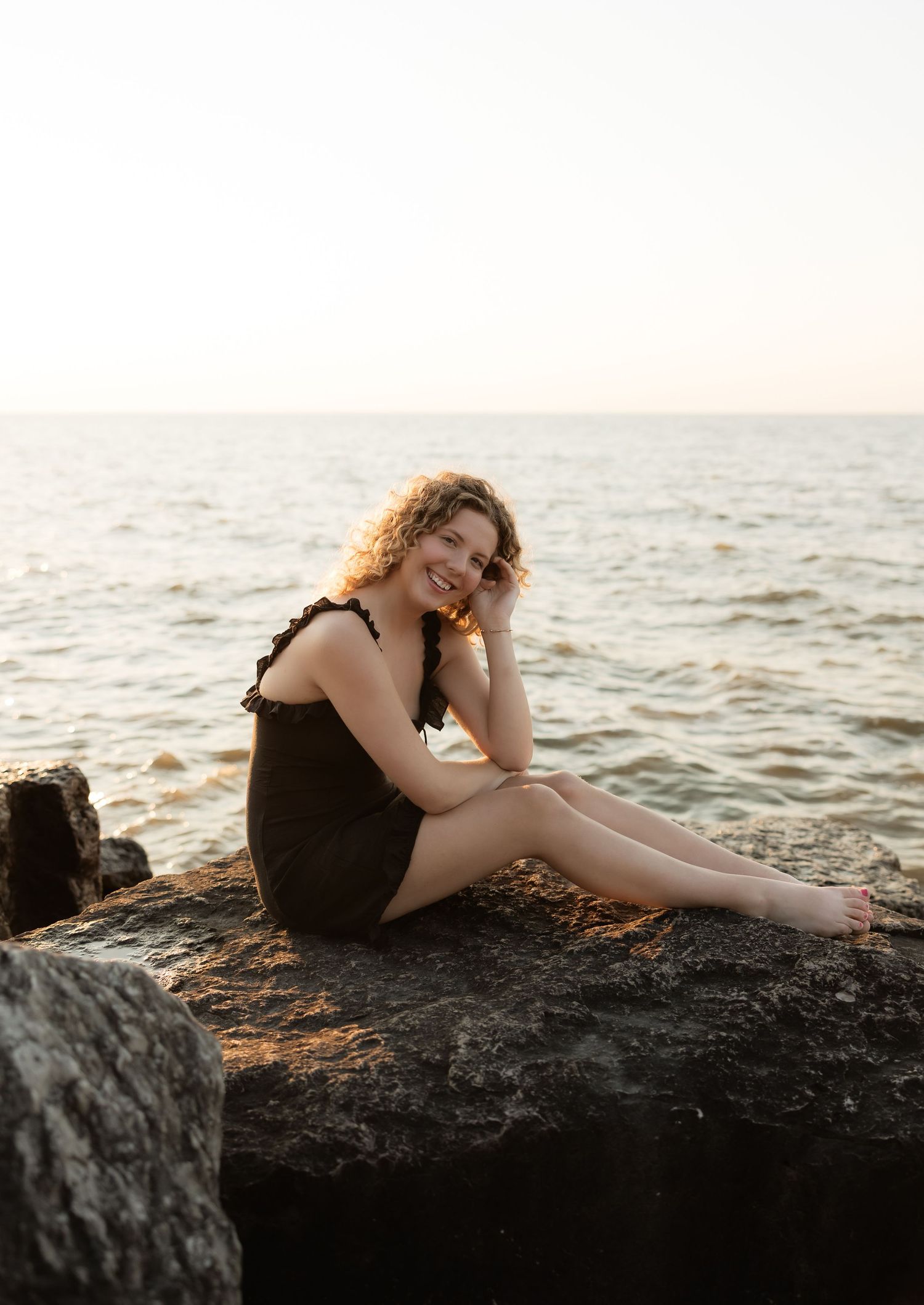 Happy curly-haired woman in black dress sitting barefoot on rocks by the sea during golden hour.