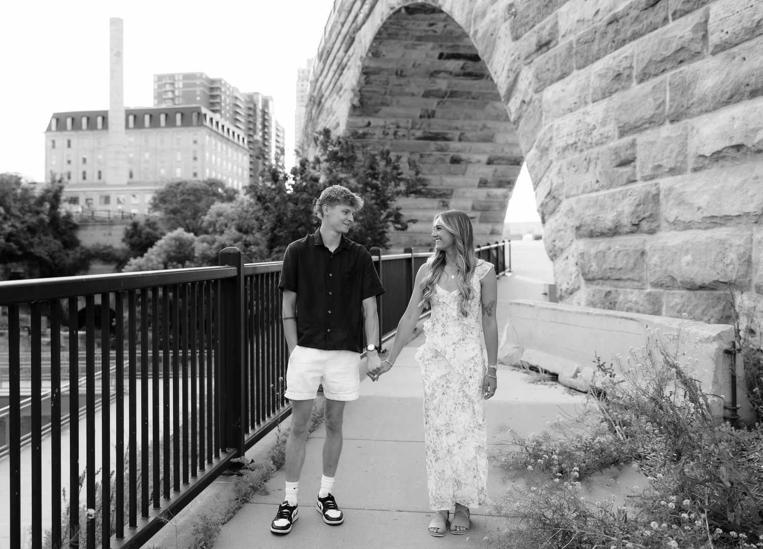 Black and white photo of a couple holding hands while walking near a stone arch bridge.